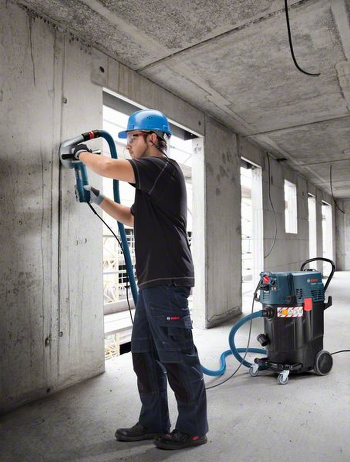 Uomo con casco blu fora muro in cemento, aspirapolvere industriale vicino.
