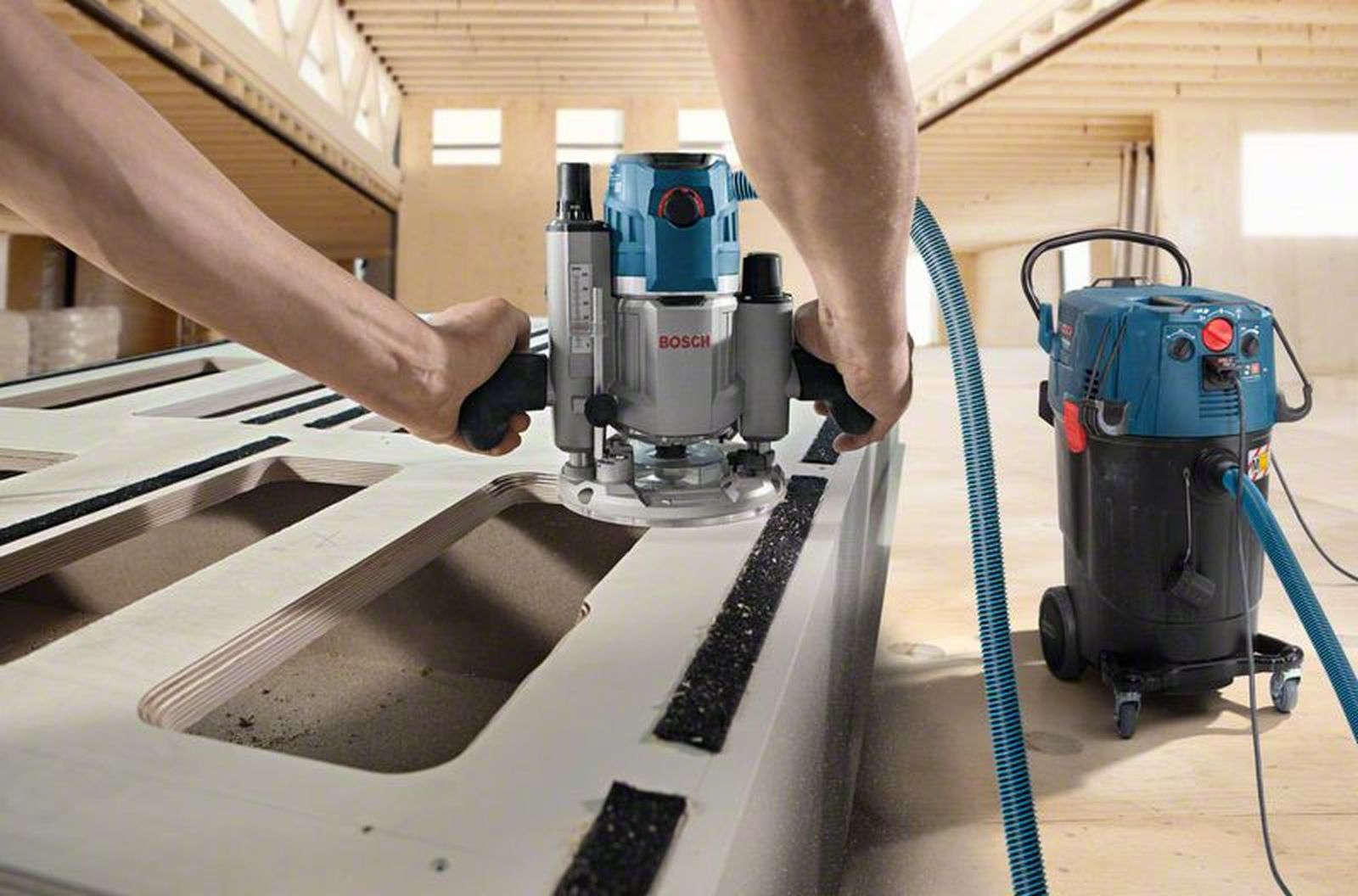 Mani usano un router Bosch su legno con un aspirapolvere nelle vicinanze.