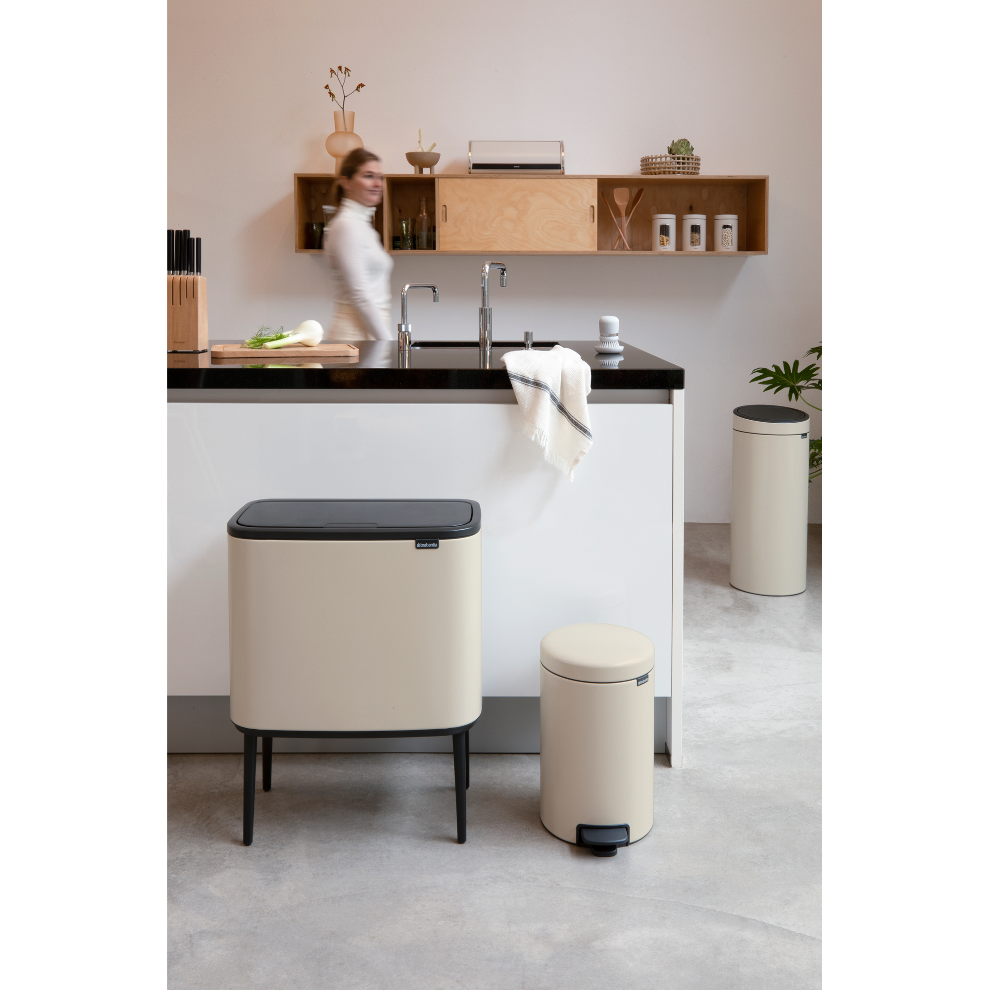 A woman in light clothing stands in a modern kitchen with beige trash cans.