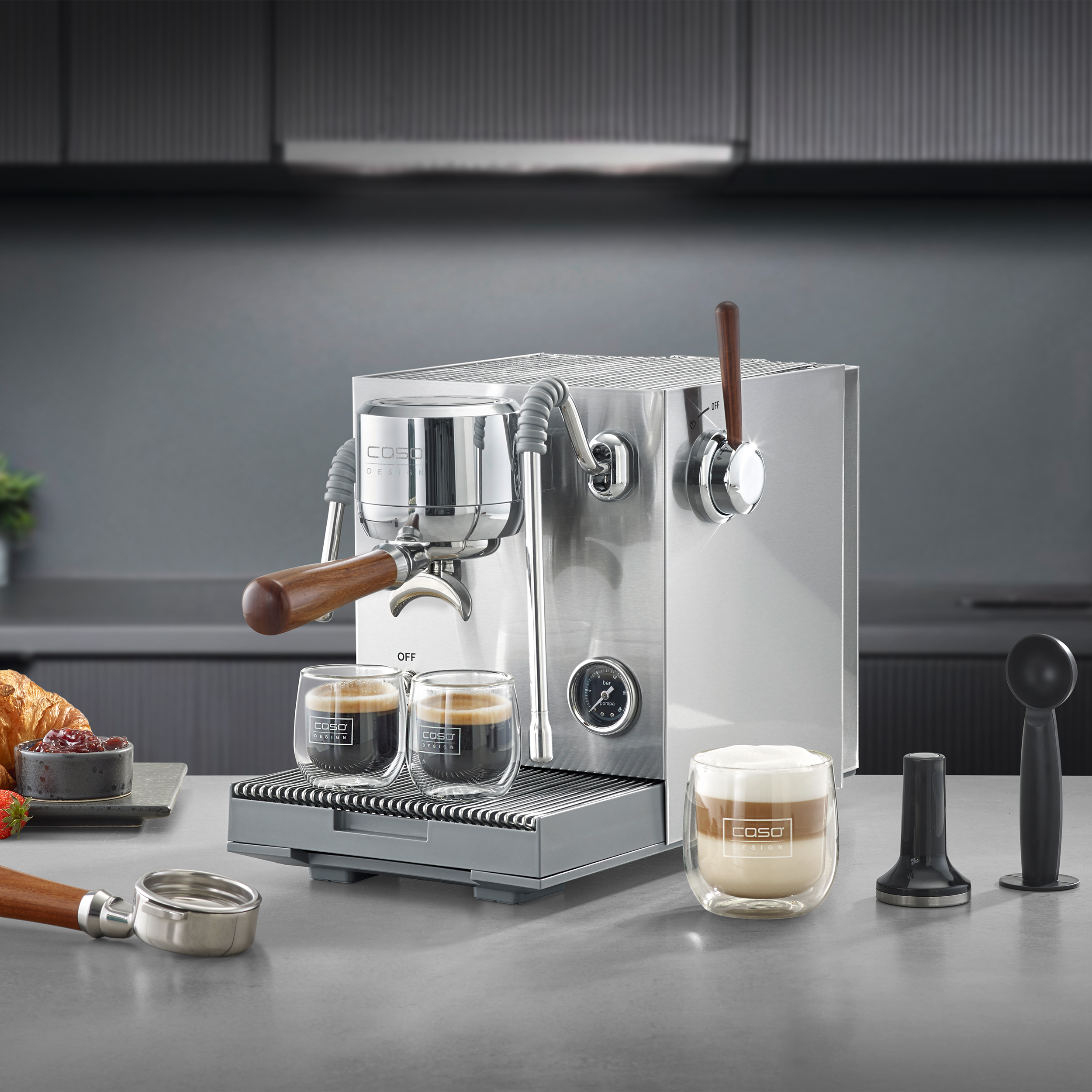 Eine silberne Espressomaschine mit Holzdetails auf einer Arbeitsplatte, daneben Kaffee und Croissant.