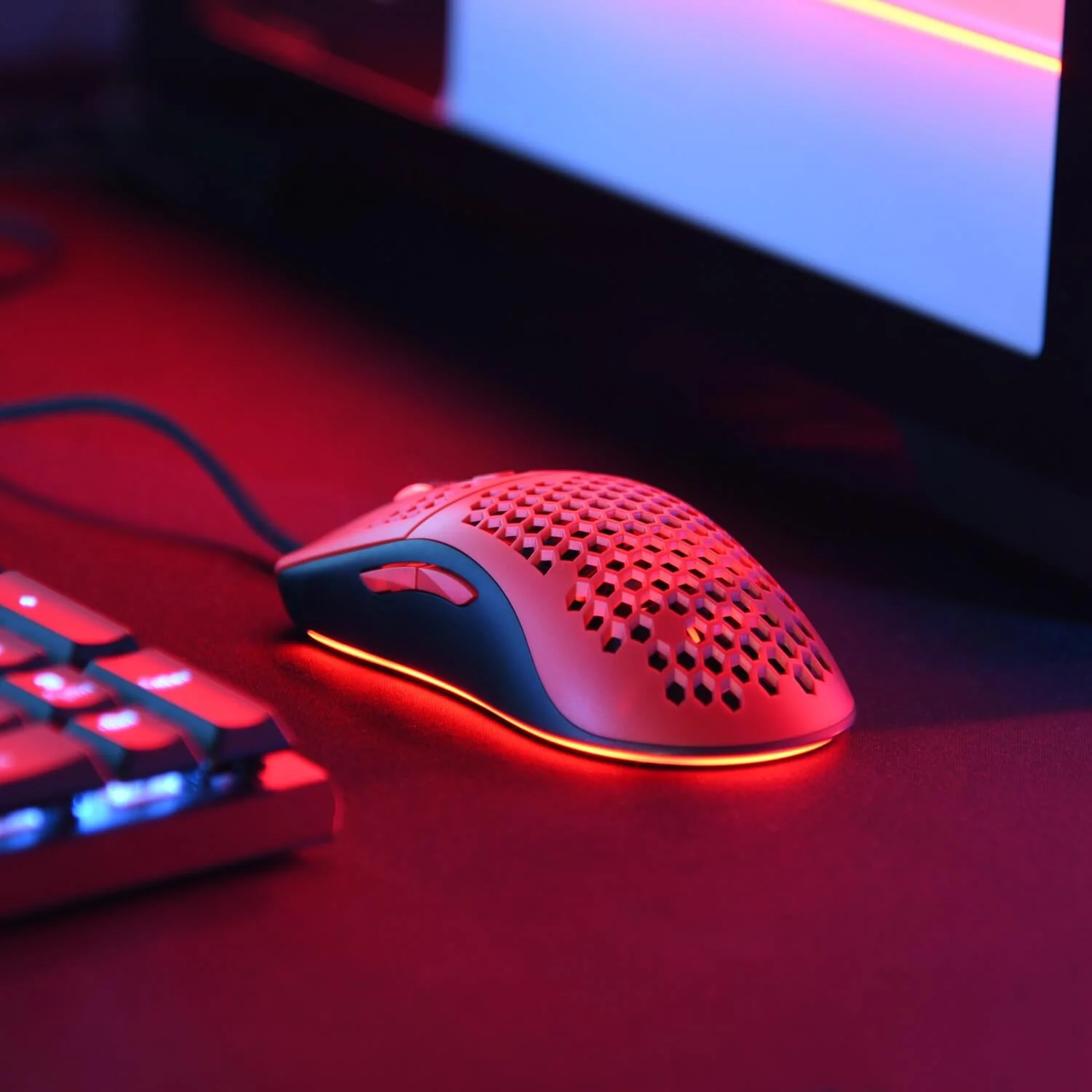 Een rode gamingmuis met honingraatpatroon licht op een bureau naast een toetsenbord.