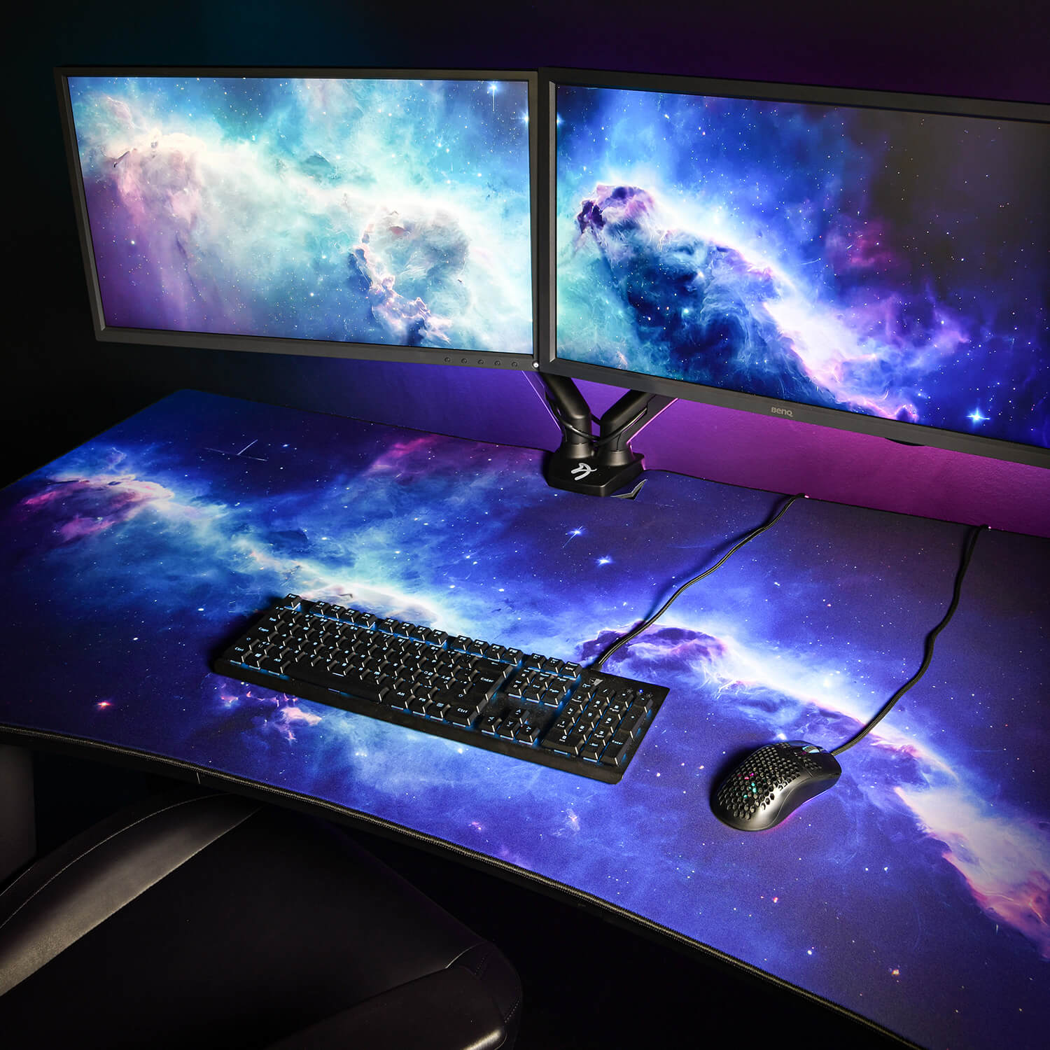 Twee monitors met sterrenstelsels, toetsenbord en muis op een bureau.