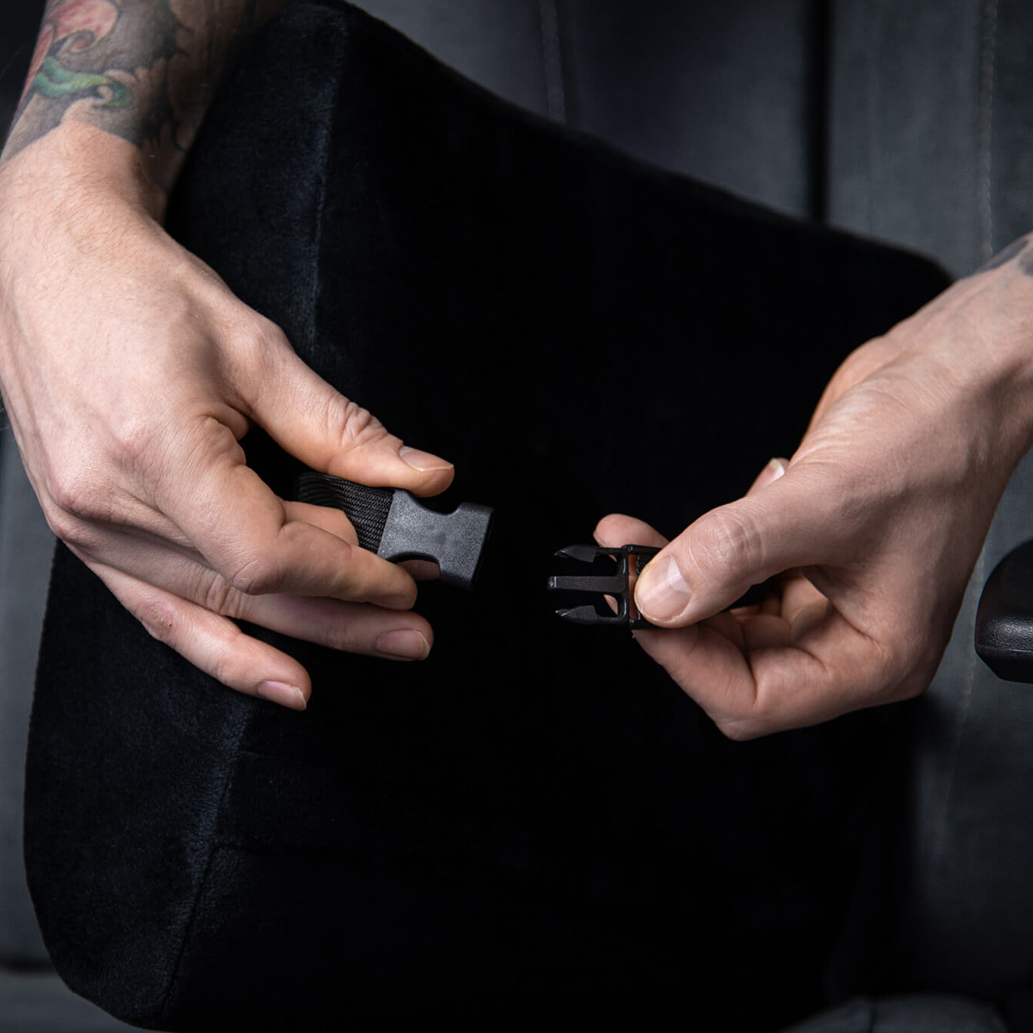 Mani che allacciano una fibbia nera su un cuscino nero. Tatuaggi visibili sul braccio.