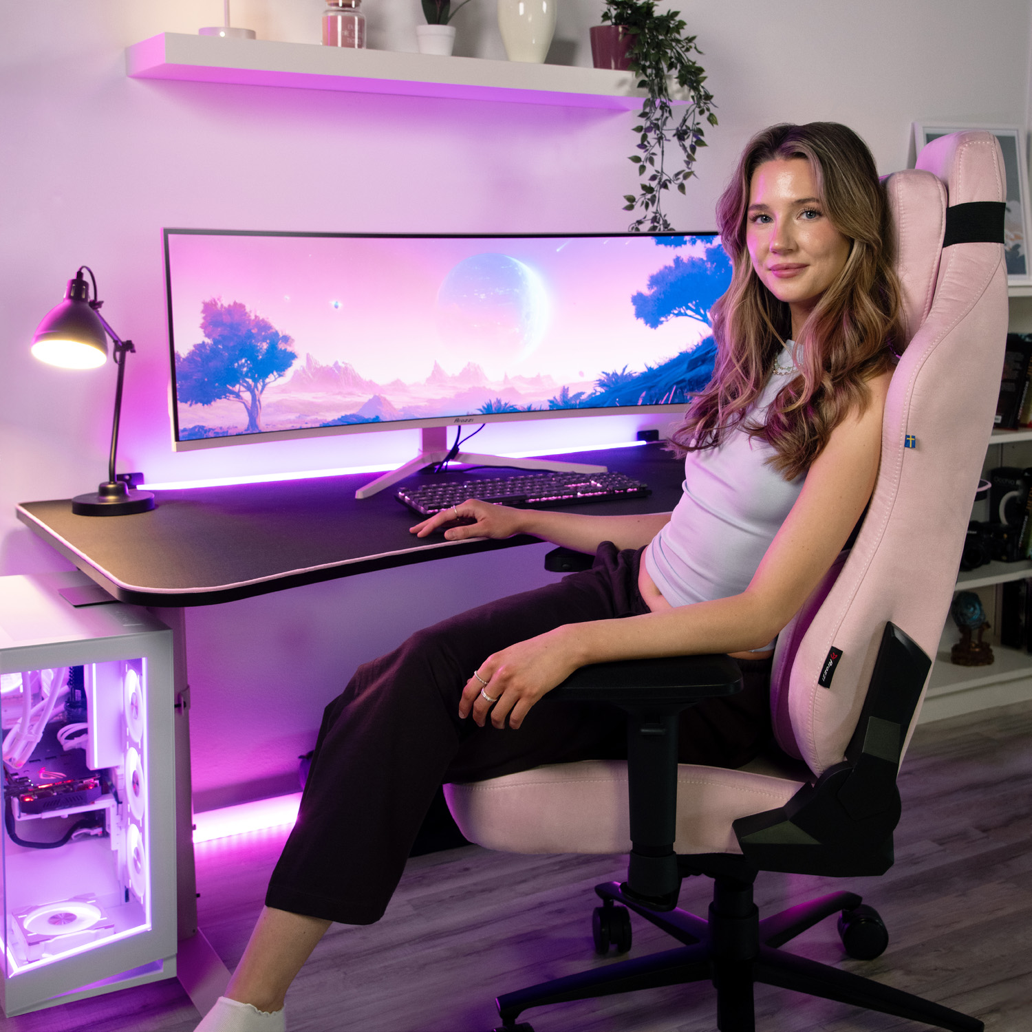 Eine junge Frau sitzt in einem rosafarbenen Gaming-Stuhl vor einem Schreibtisch mit einem großen Monitor.
