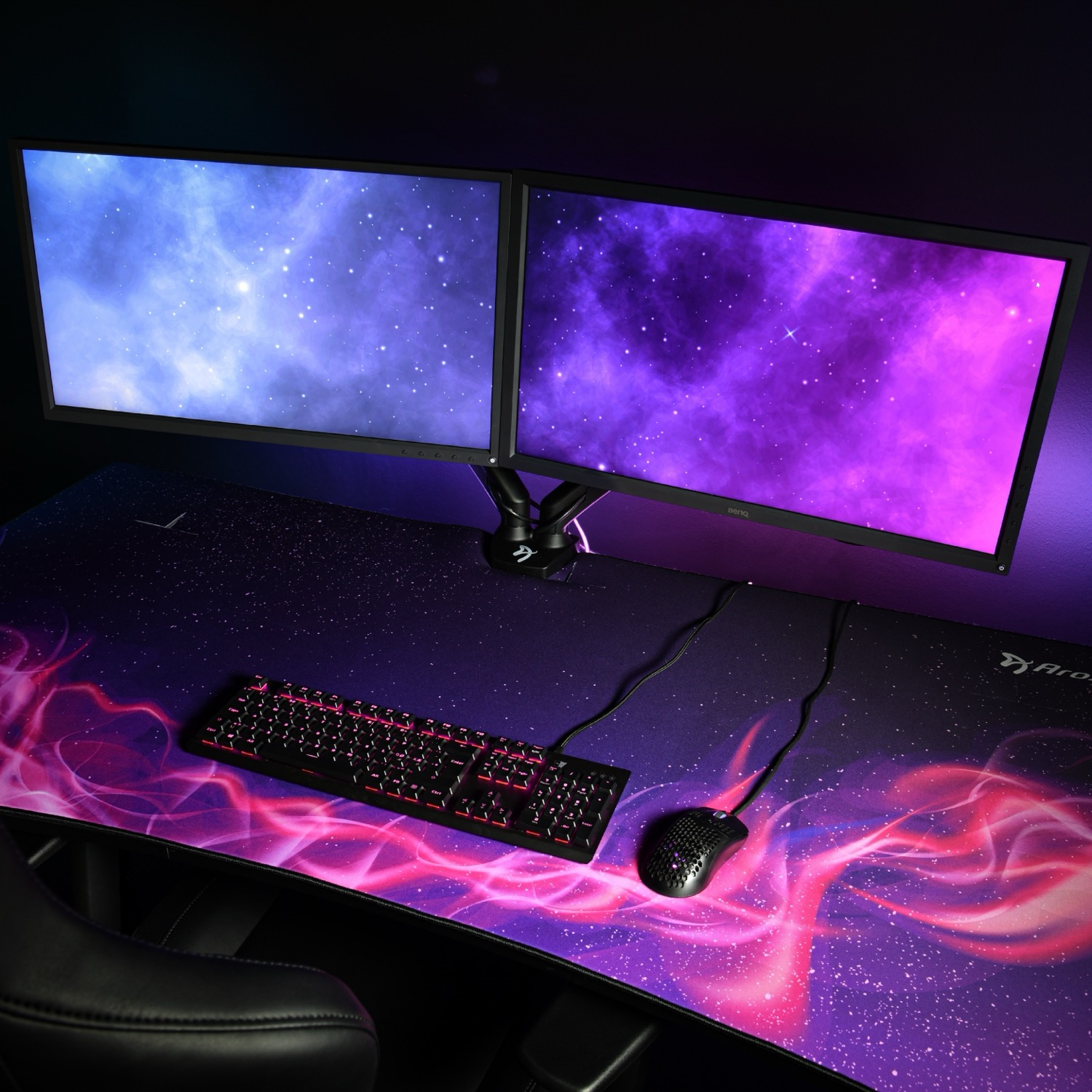Ein Dual-Monitor-Setup mit Galaxie-Hintergründen, einer beleuchteten Tastatur und Maus auf einer Schreibtischunterlage.