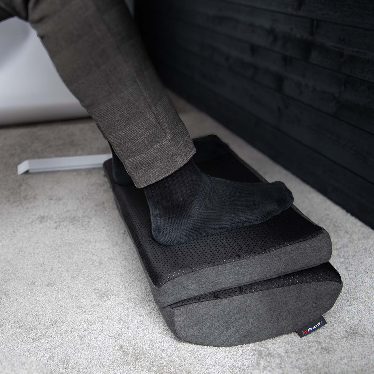 Una persona con pantaloni grigi a quadri e calzini neri appoggia un piede su un poggiapiedi nero.