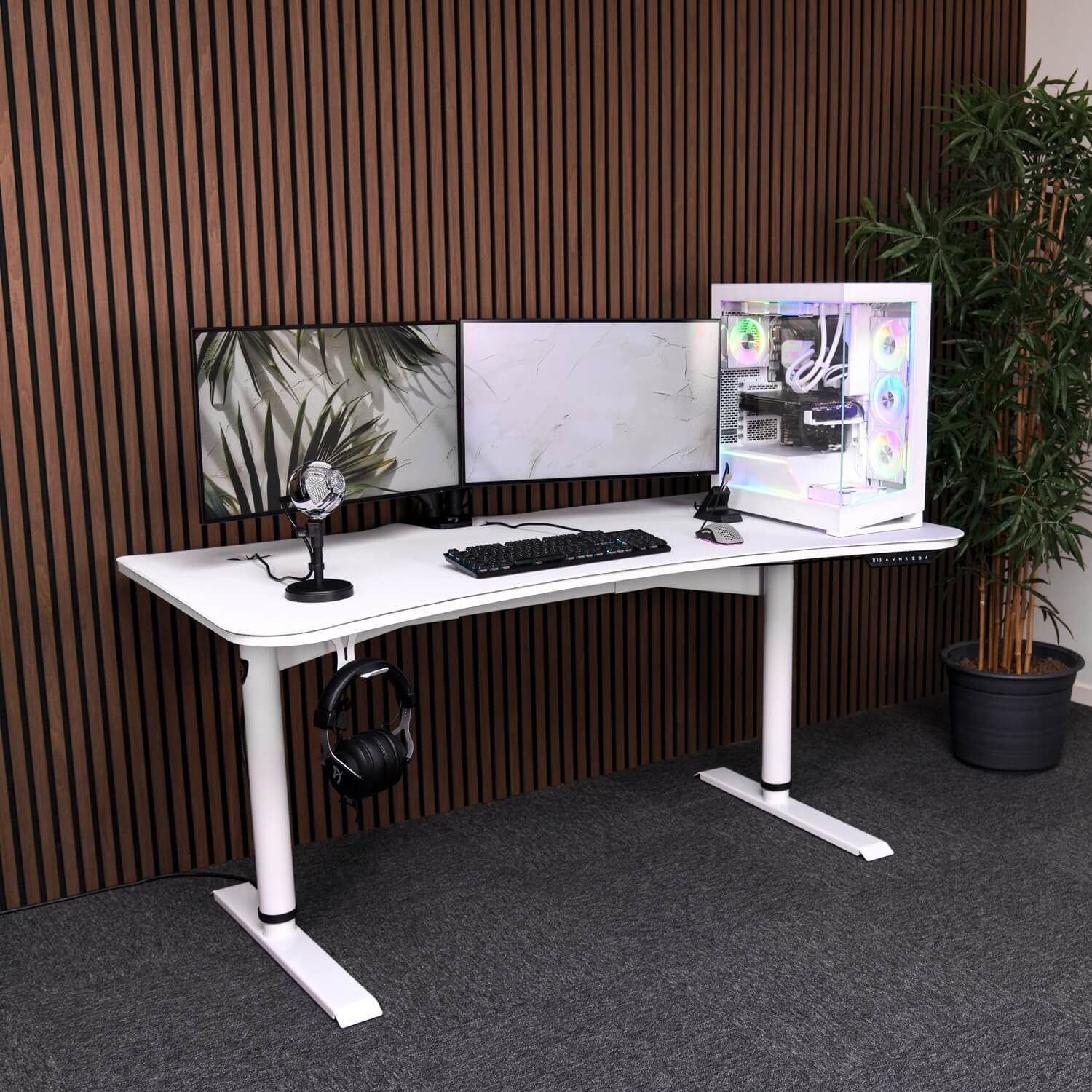 Een wit bureau met drie monitoren, pc, toetsenbord, muis, microfoon en koptelefoon. Rechts een plant.