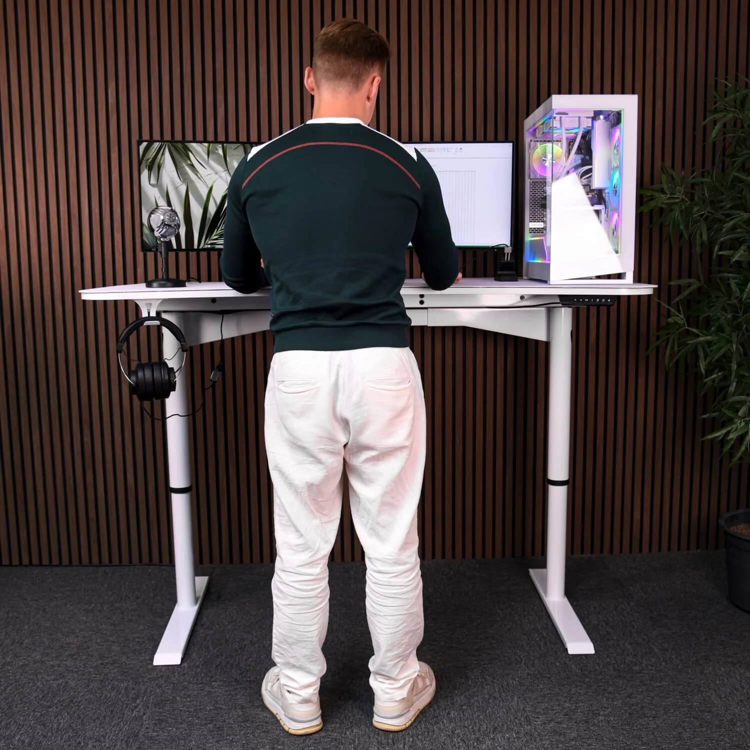 Man staat aan een wit bureau met computer, monitor en microfoon.