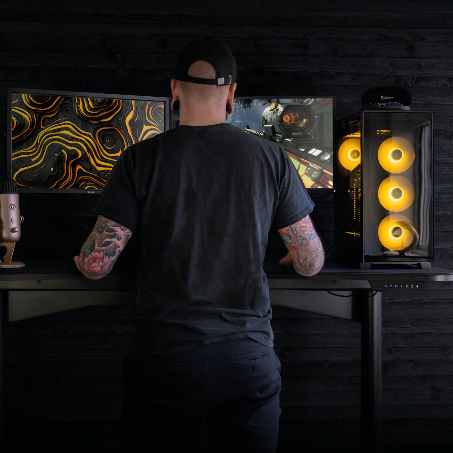 Uomo tatuato gioca ai videogiochi su due monitor, accanto a un PC da gaming con luci gialle.