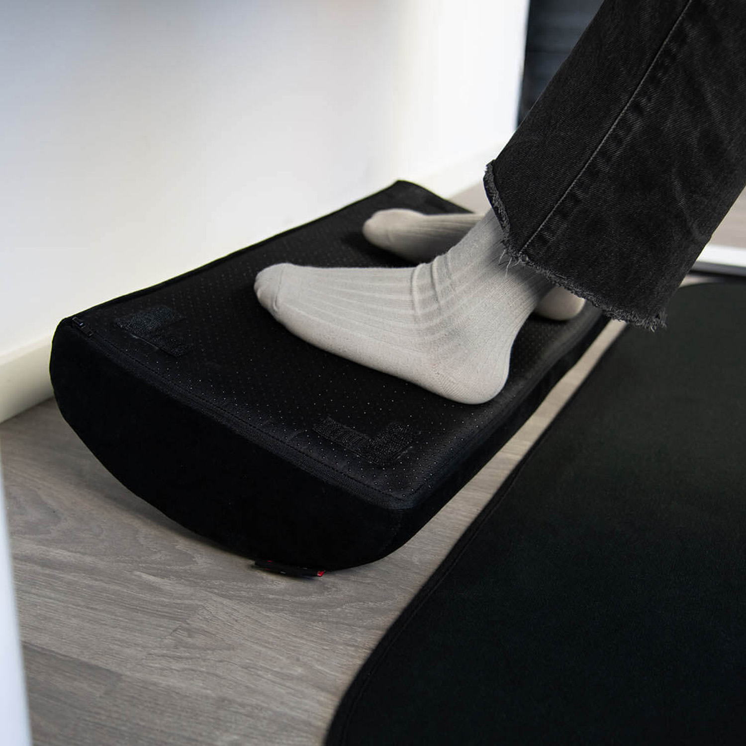 Una persona con calzini grigi e jeans neri sta su un poggiapiedi nero su pavimento in legno.