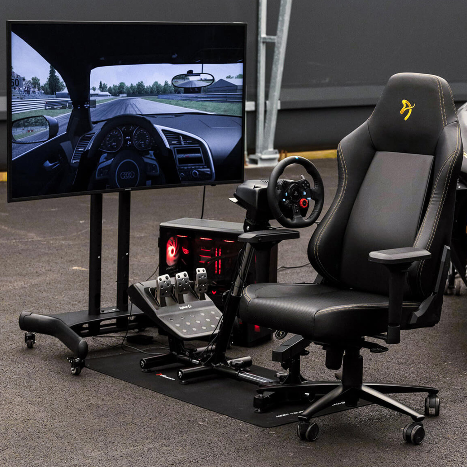 Una configuración de simulador de carreras con pantalla grande que muestra el interior de un coche en una pista, silla de carreras negra.