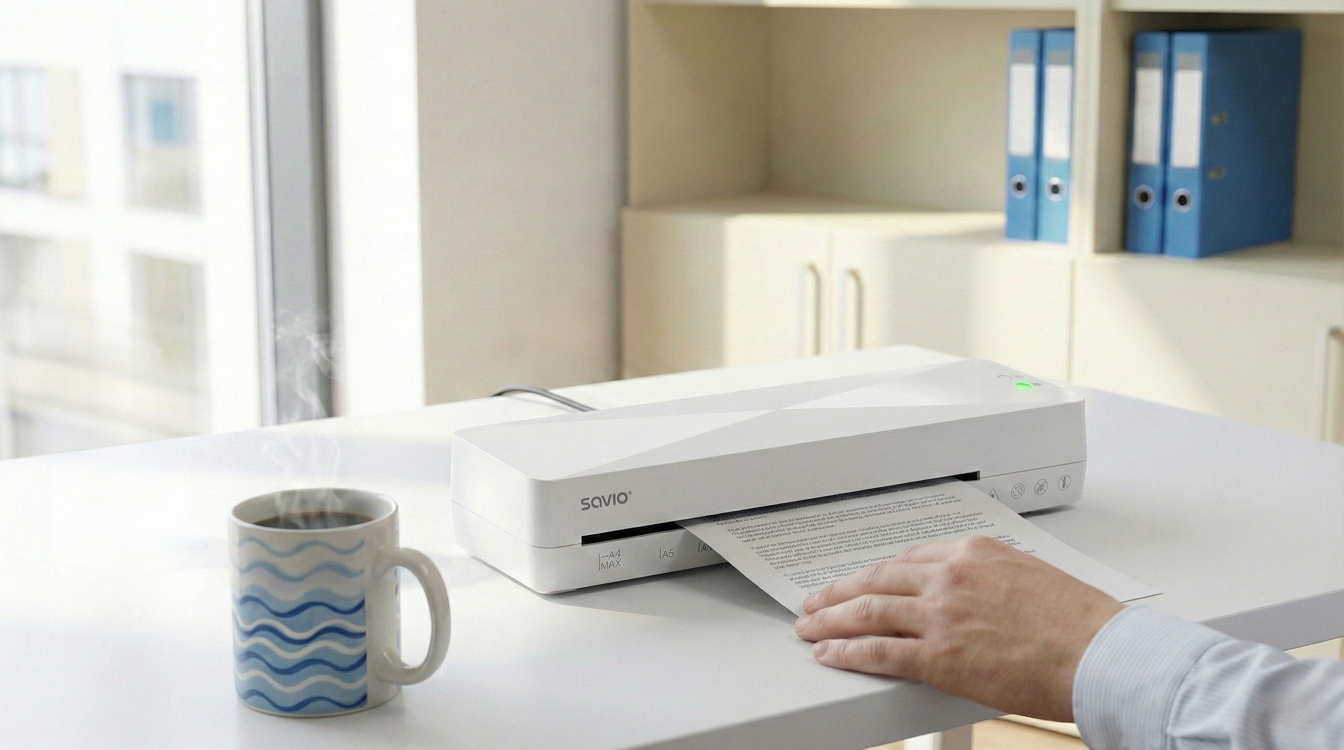 Biała laminarka Savio na biurku z parującą filiżanką kawy obok.