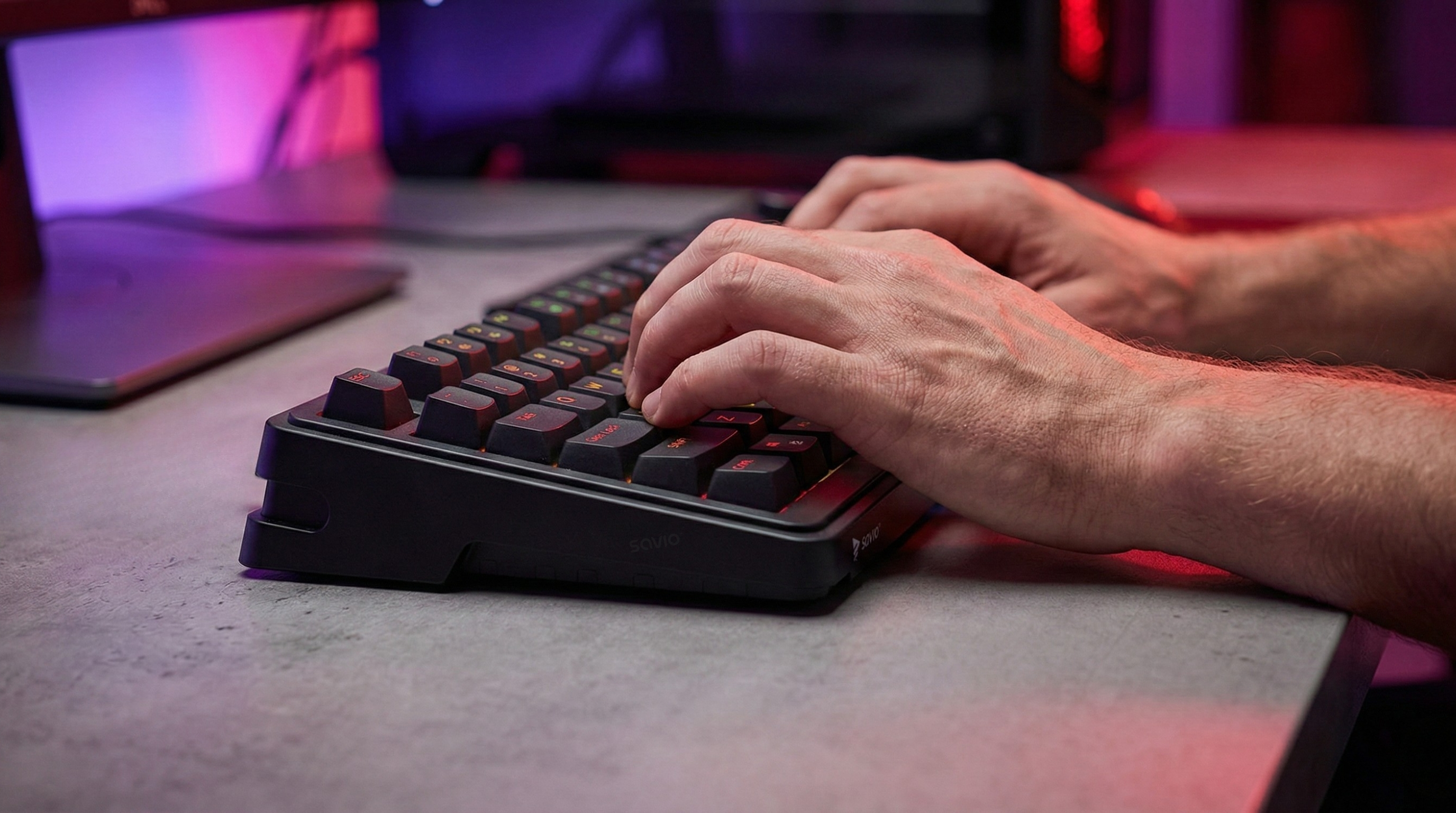 Dłonie piszące na czarnej klawiaturze z czerwonymi akcentami, fioletowe i różowe światło w tle.