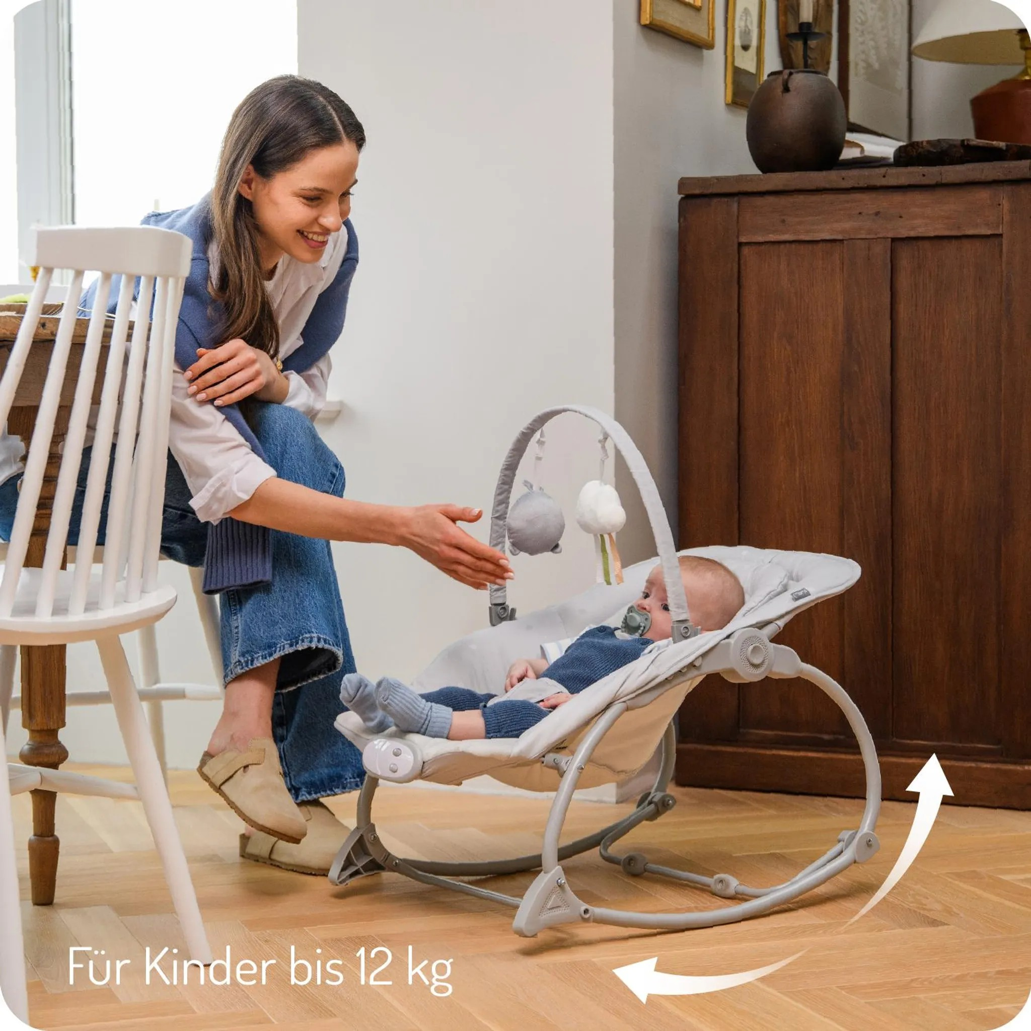 Eine Frau sitzt an einem Tisch und lächelt ein Baby in einer Babywippe an.