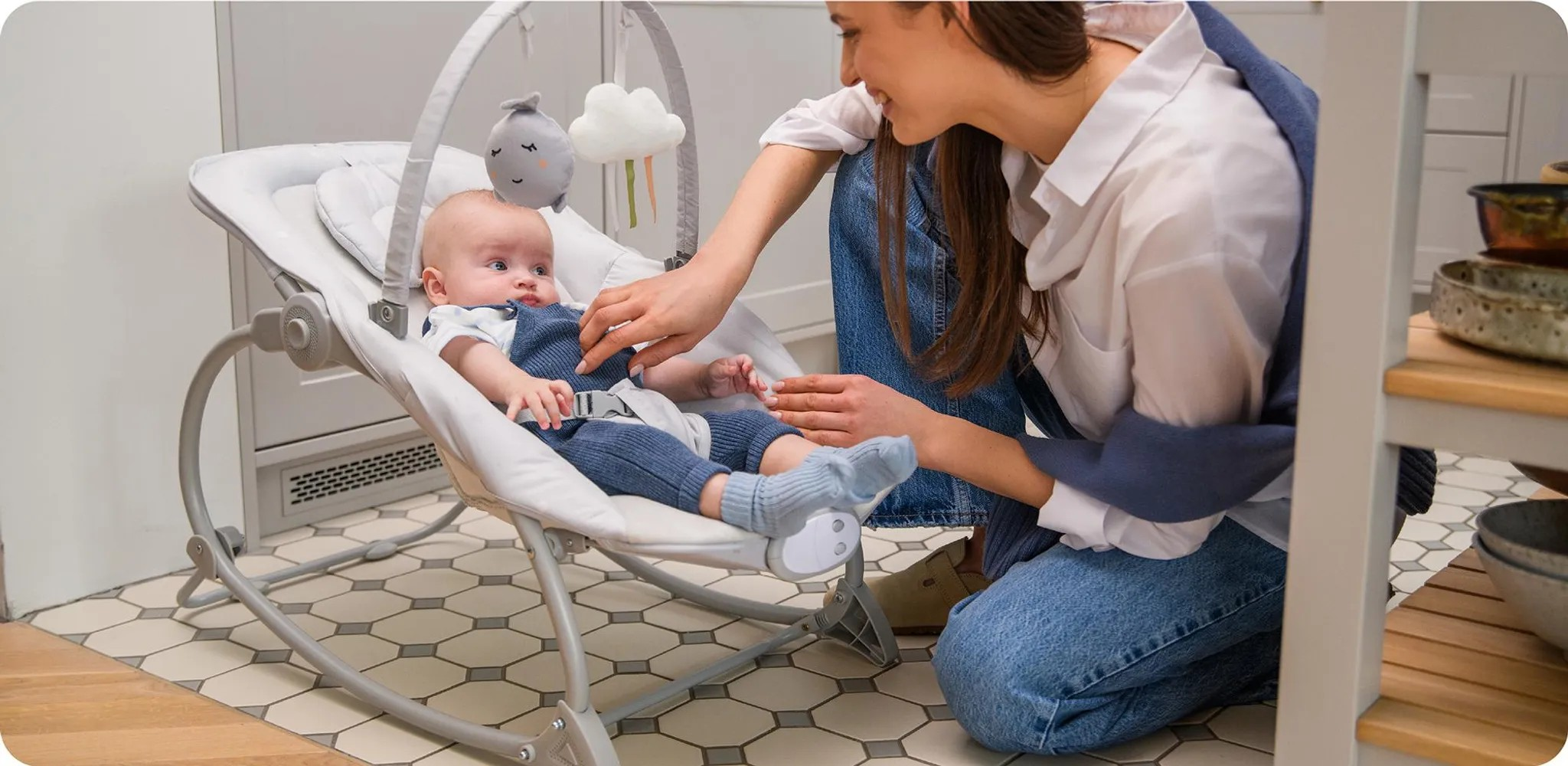 Eine Frau kniet neben einem Baby in einer Wippe. Spielzeug hängt über dem Baby.