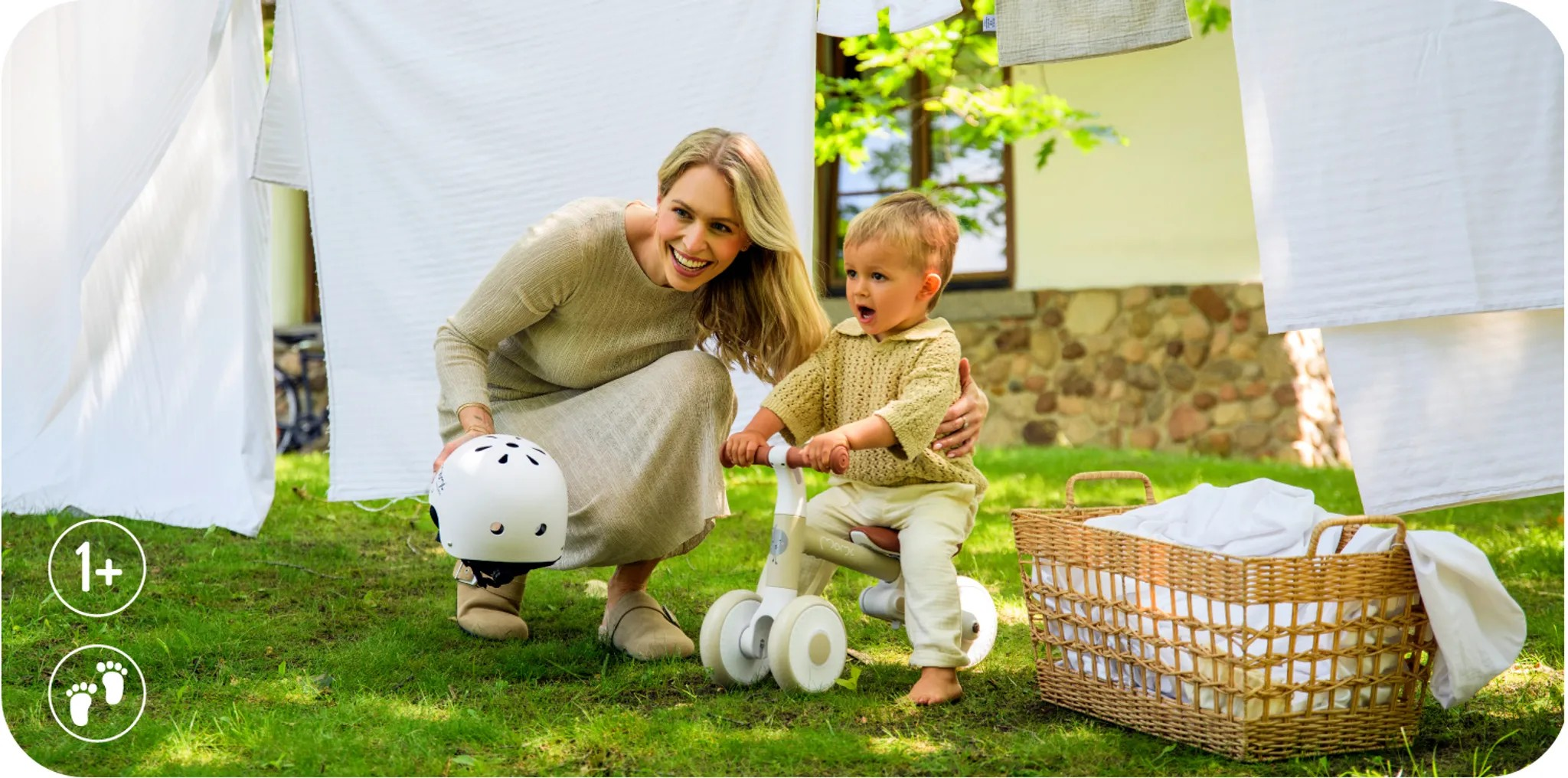 Eine Frau und ein Kind mit einem Laufrad im Gras, Wäsche hängt im Hintergrund.