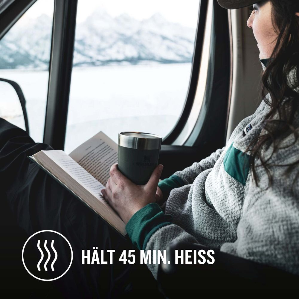 Una persona legge un libro tenendo una tazza in un veicolo con vista sulle montagne.