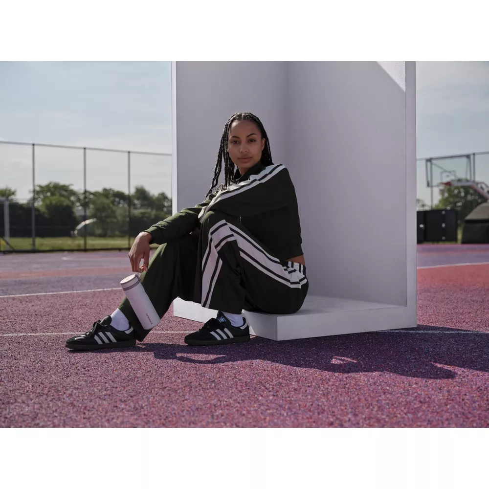 Mujer con chándal Adidas verde oscuro sentada en repisa blanca, sostiene botella agua.