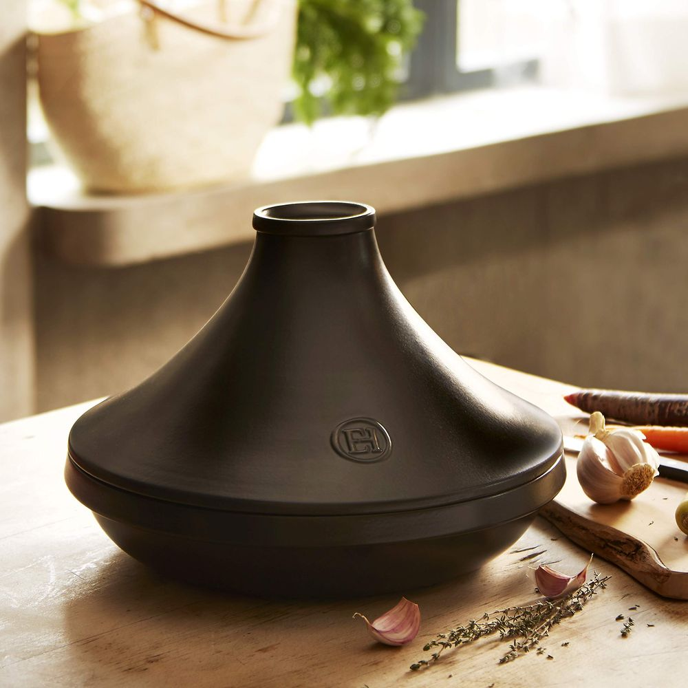 Ein schwarzer Tajine-Topf steht auf einem Holztisch mit Knoblauch und Kräutern.