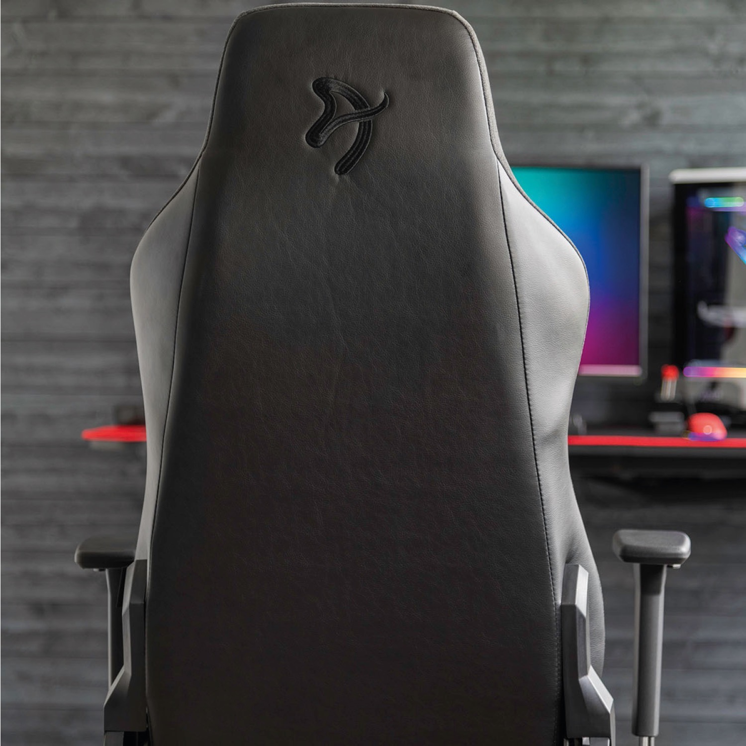 Vista posteriore sedia gaming nera con logo. Monitor e PC visibili.