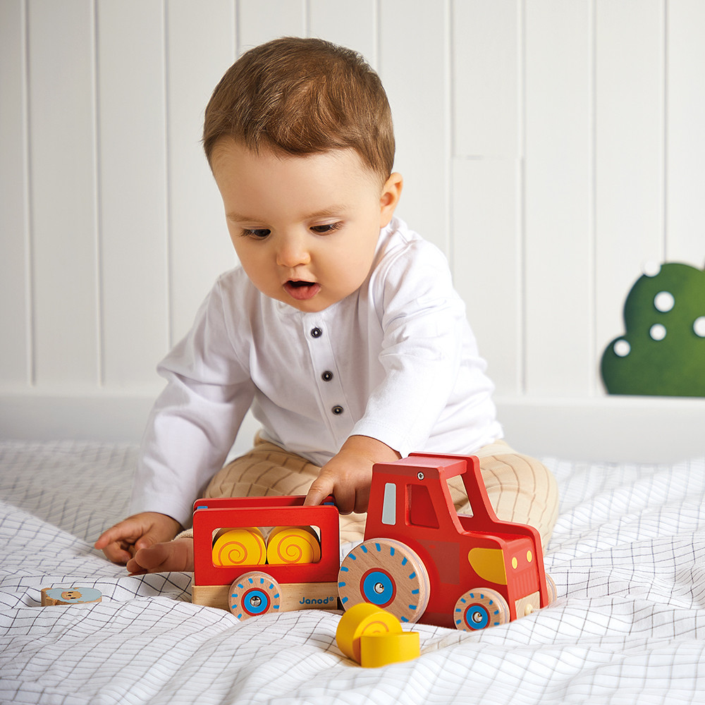 Ein Baby in einem weißen Hemd spielt mit einem roten Holzschlepper und Anhänger auf einer karierten Decke.