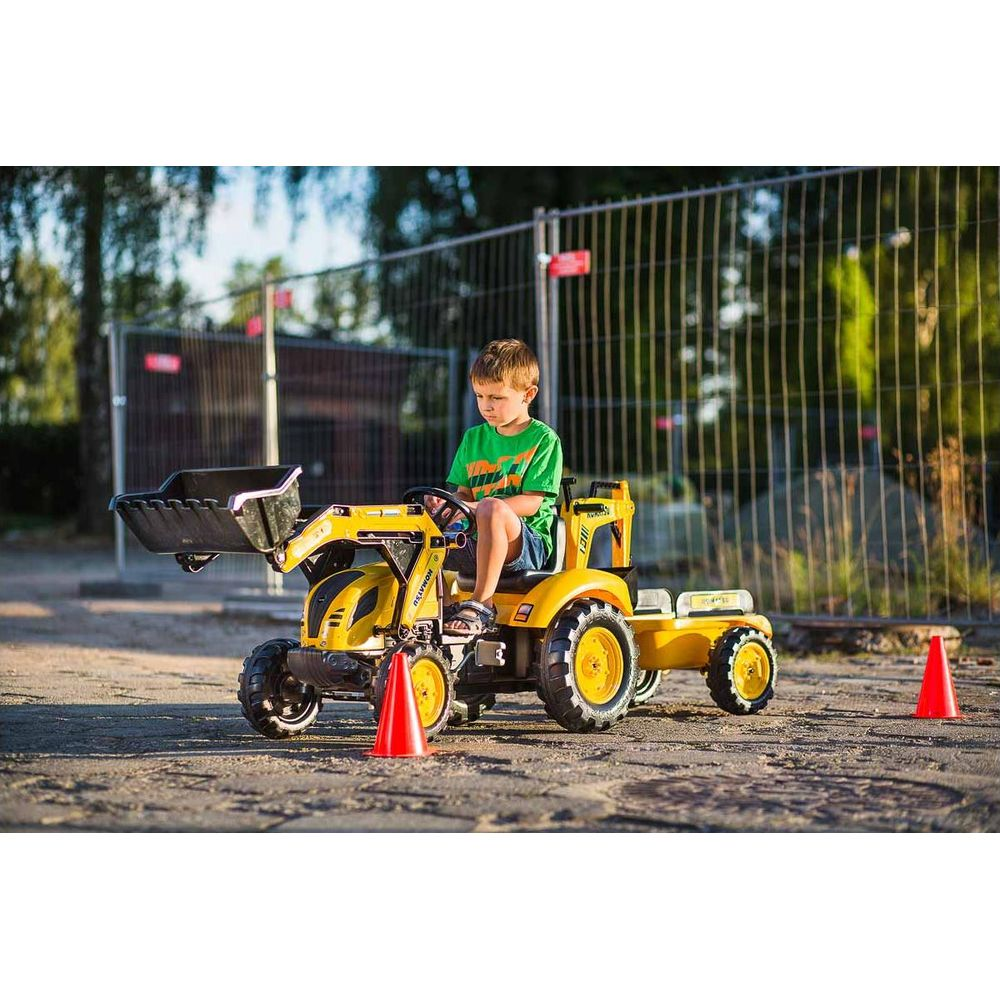 Ein Junge fährt einen gelben Spielzeugtraktor mit Lader und Anhänger. Orangenkegel sind in der Nähe.