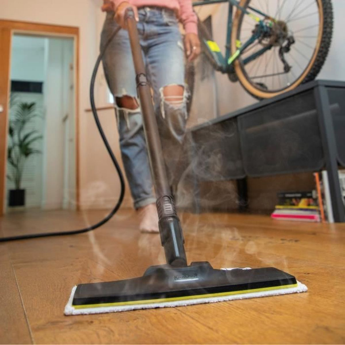 Pulitore a vapore su pavimento in legno. Persona in jeans e top rosa. Bicicletta sullo sfondo.