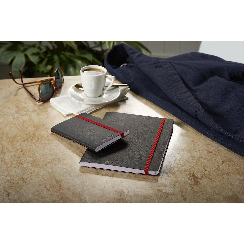 Zwei Notizbücher mit rotem Gummiband, eine Kaffeetasse, Sonnenbrille und Jacke liegen auf einem Tisch.