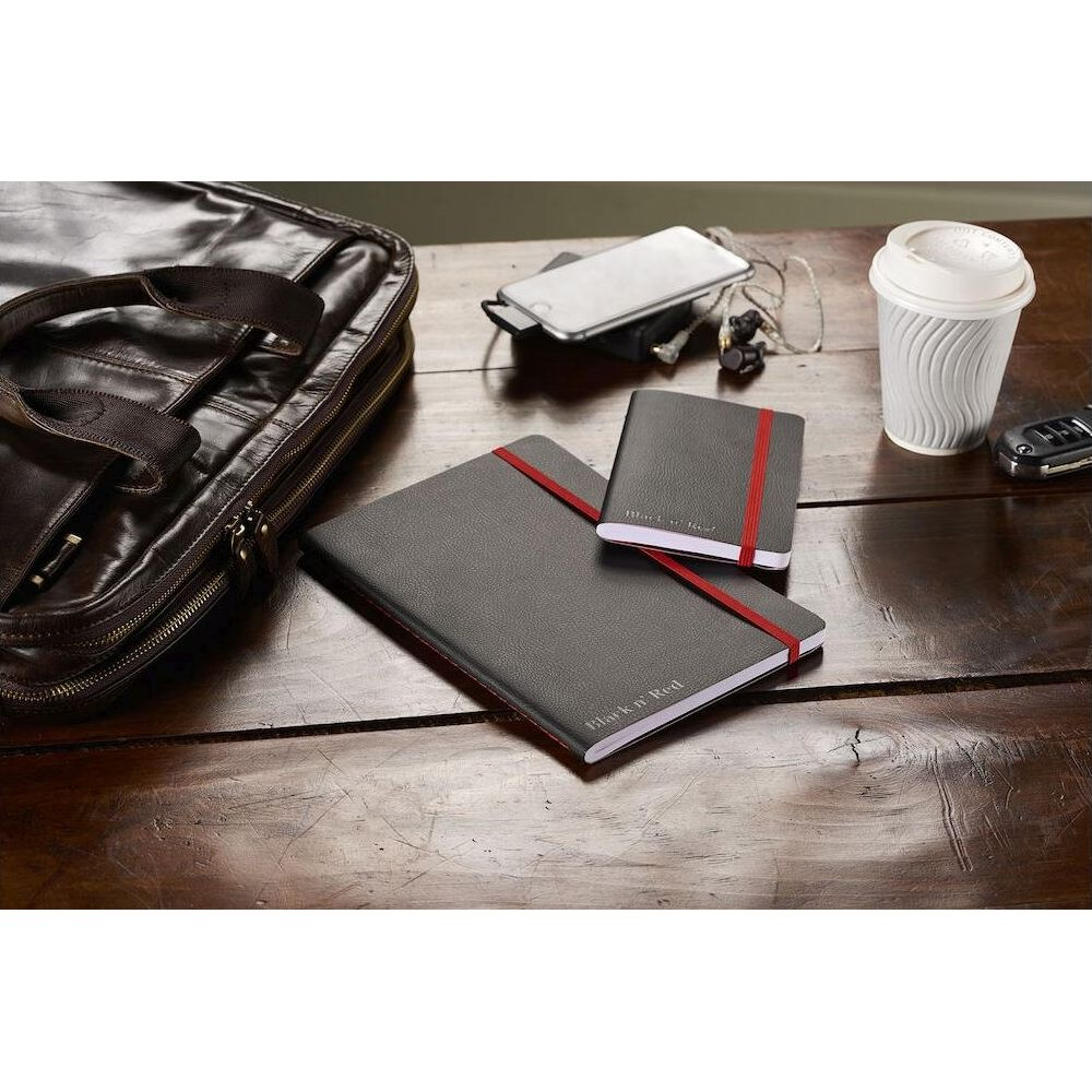 Eine dunkle Ledertasche, zwei Notizbücher mit rotem Band, ein Handy, Kopfhörer, ein Becher und ein Schlüssel.
