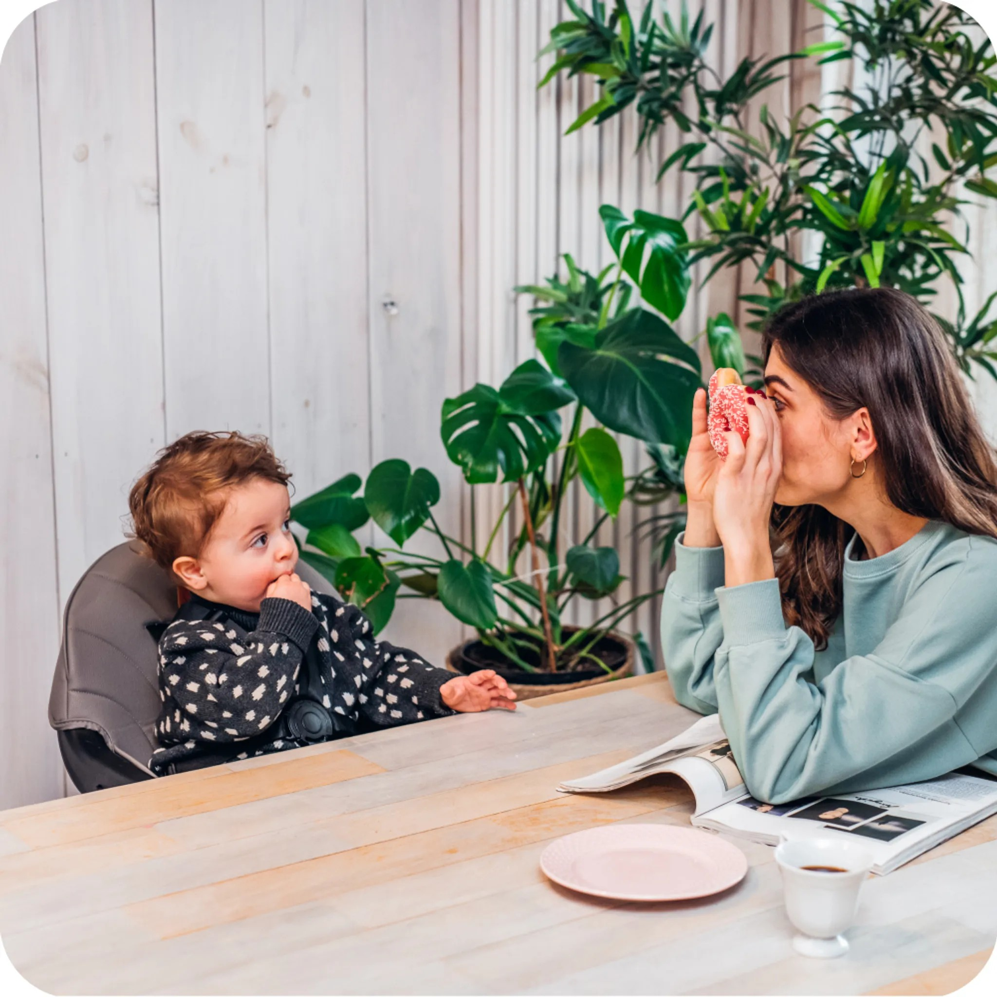 Eine Frau hält einen Donut vor ihr Gesicht, ein Baby sitzt im Hochstuhl daneben.