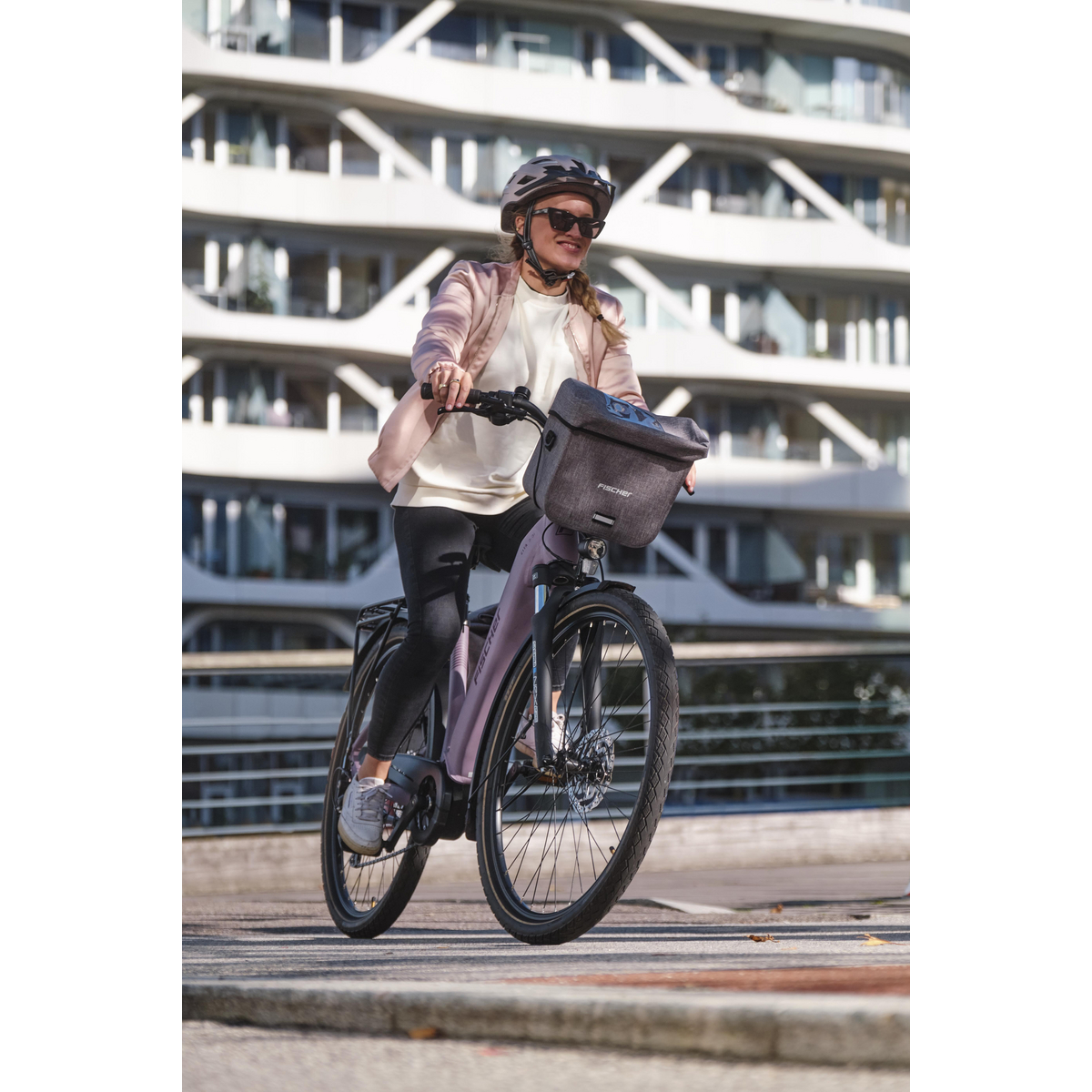Eine Frau mit Helm fährt ein pinkes Fahrrad mit Lenkertasche vor einem modernen Gebäude.