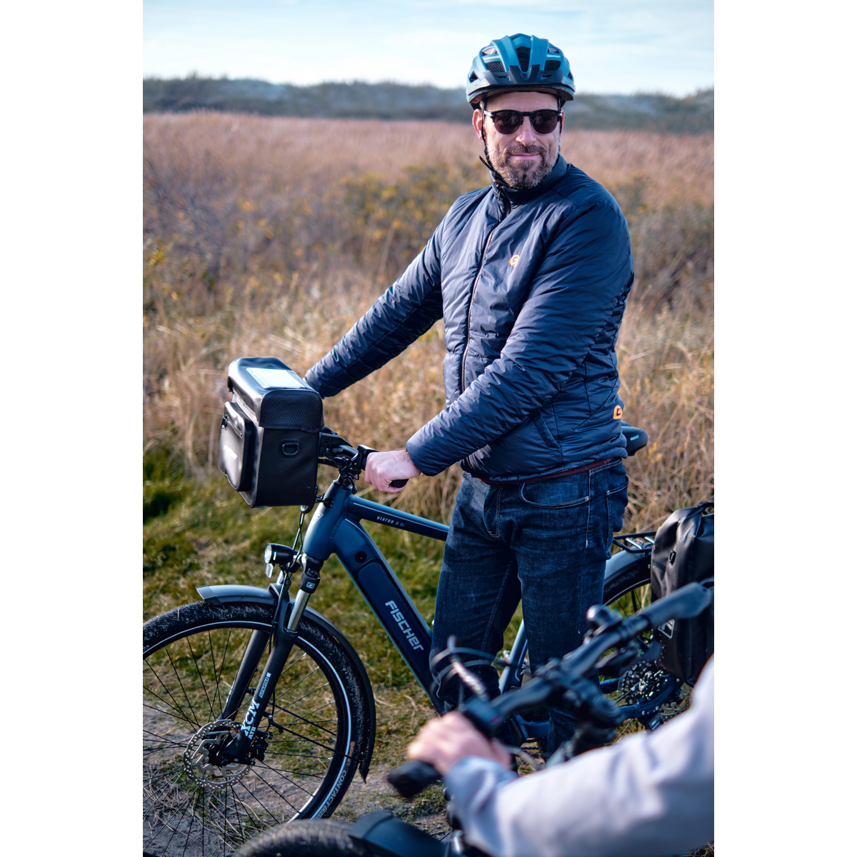 Mann mit Helm und Sonnenbrille steht neben blauem Fahrrad mit Lenkertasche.