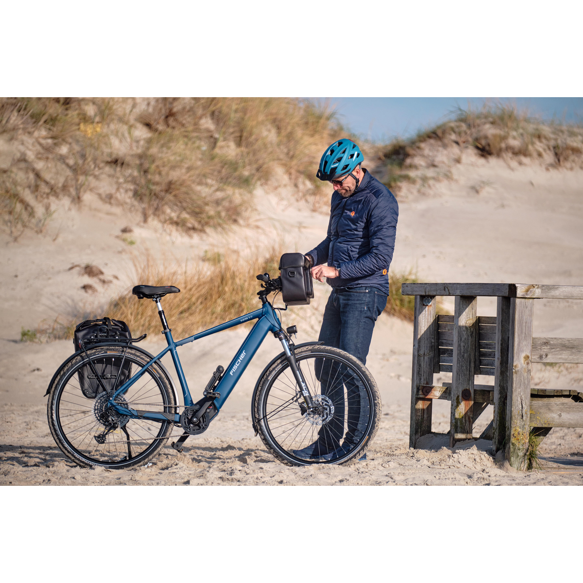 Ein Mann in blauer Jacke und Helm steht neben einem blauen Fahrrad am Strand.