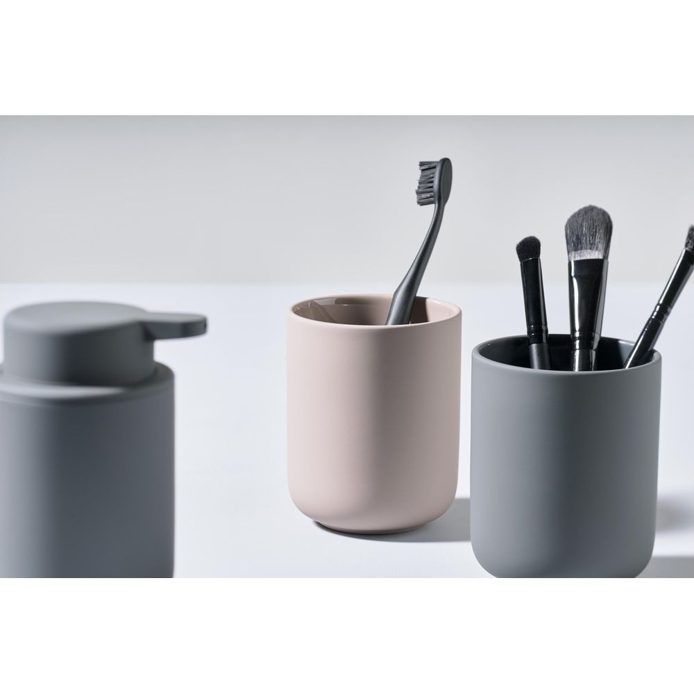 Ein grauer Seifenspender, eine rosa Tasse mit schwarzer Zahnbürste und ein grauer Becher mit Pinseln.