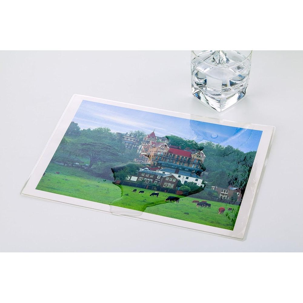 Un verre d'eau clair repose sur une feuille de plastique transparent au-dessus d'une photo de maisons et de vaches.
