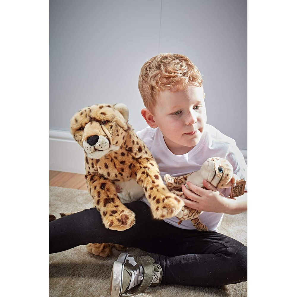 Un bambino seduto su un tappeto tiene in mano due giocattoli ghepardo di peluche. Uno grande a sinistra, uno piccolo a destra.