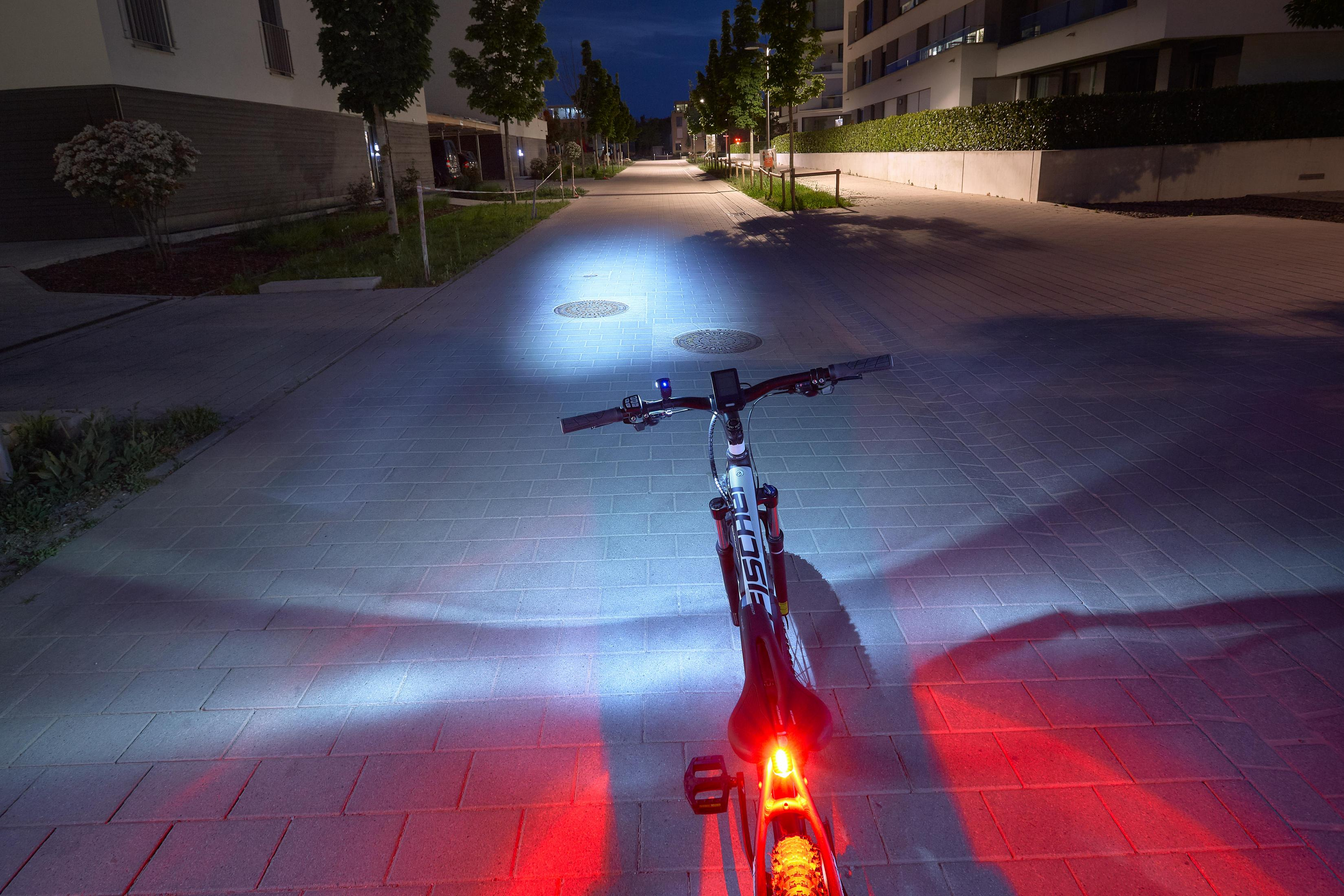 Ein Fahrrad mit Licht auf gepflastertem Weg bei Nacht, Gebäude im Hintergrund.