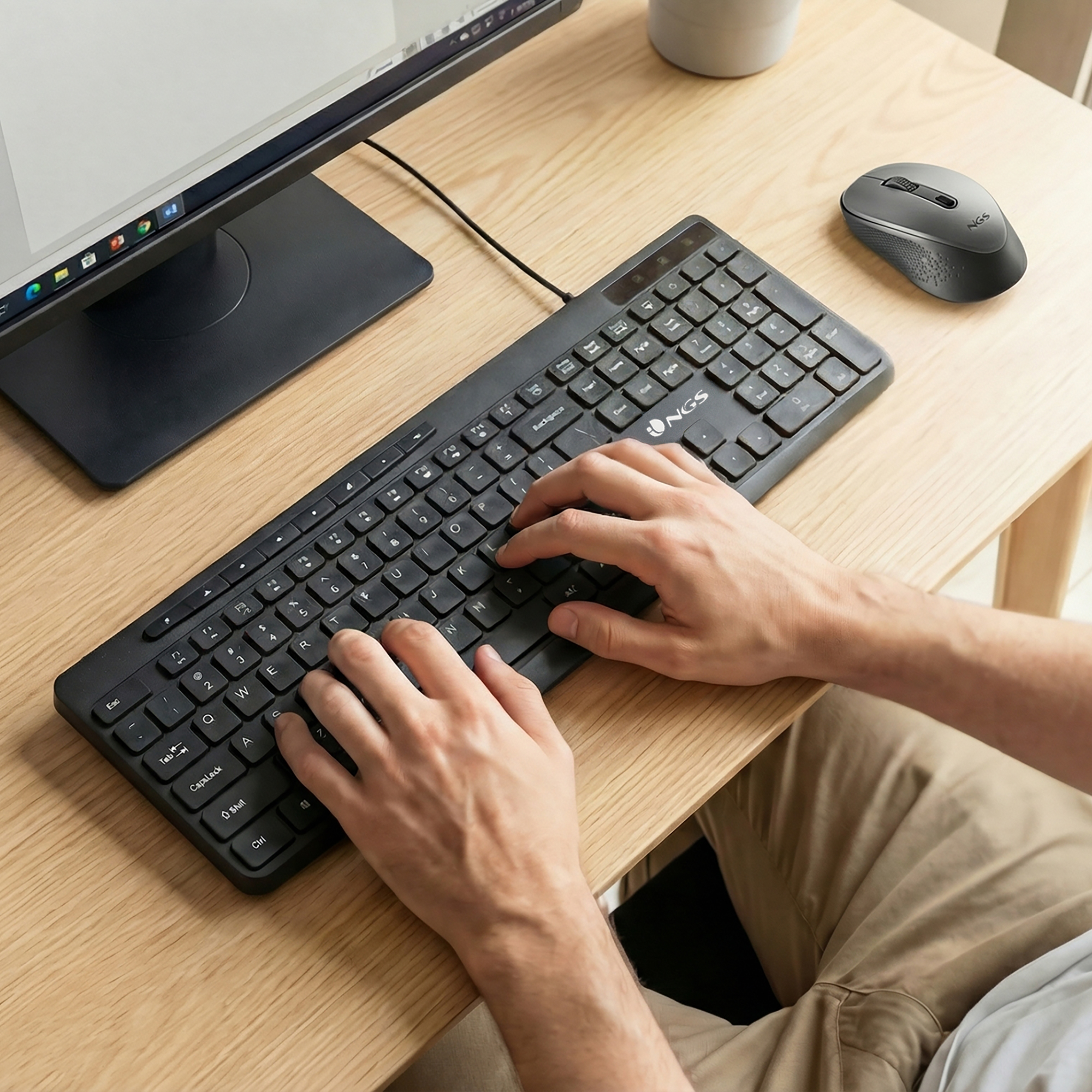 Persona digita su tastiera nera, mouse accanto, monitor su scrivania legno.