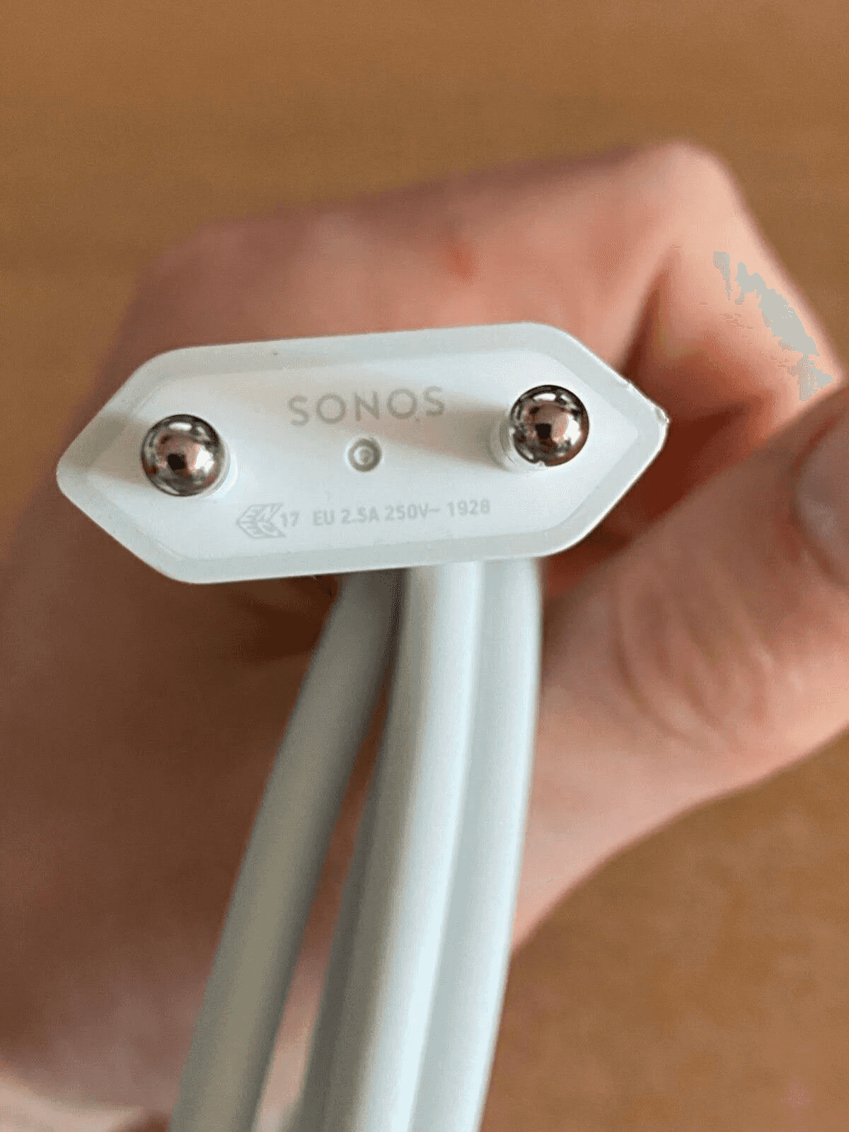 Enchufe Sonos blanco con dos pines metalicos y cables, sostenido por una mano.