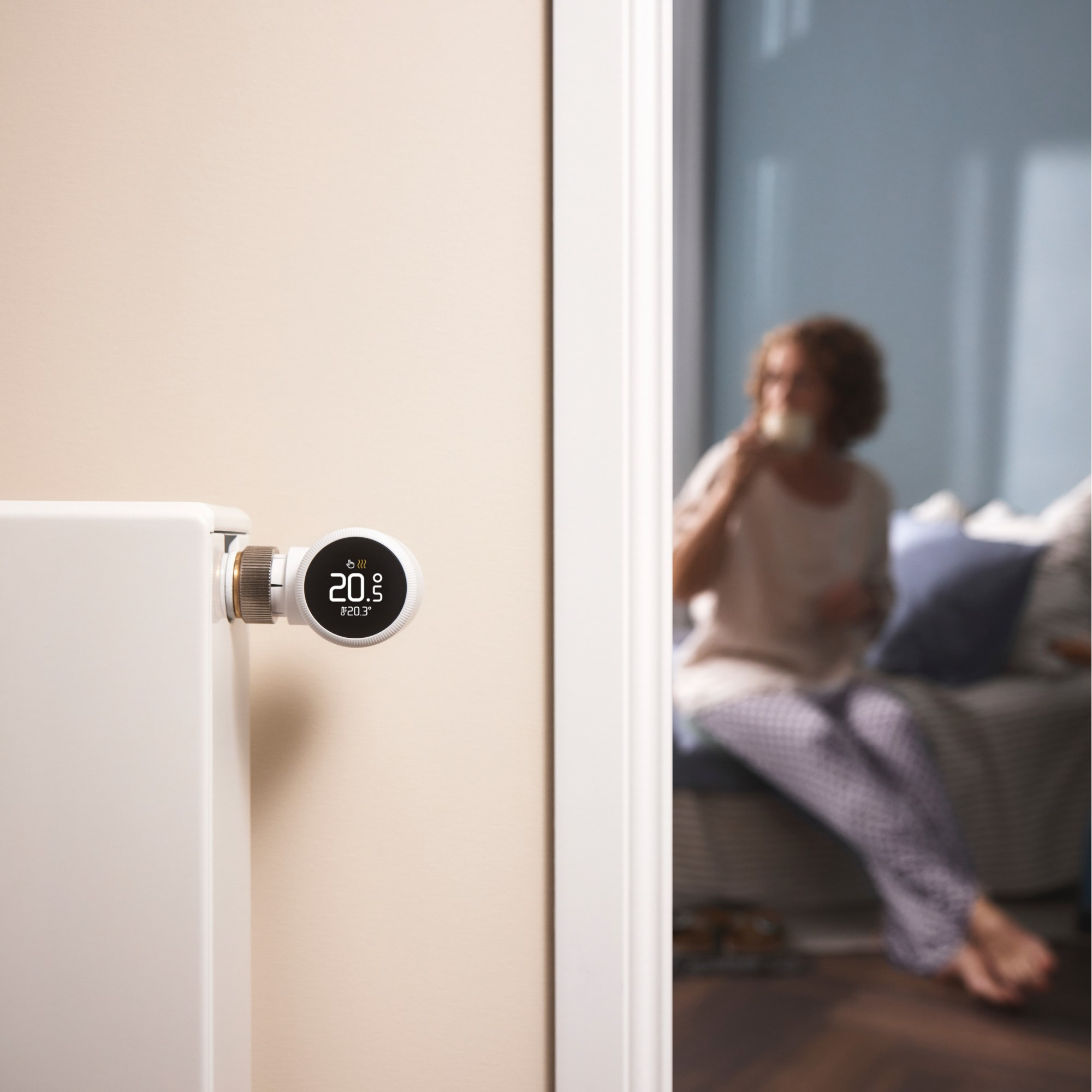 Een thermostaat toont 20,5 graden naast een radiator. Een vrouw drinkt thee op de achtergrond.