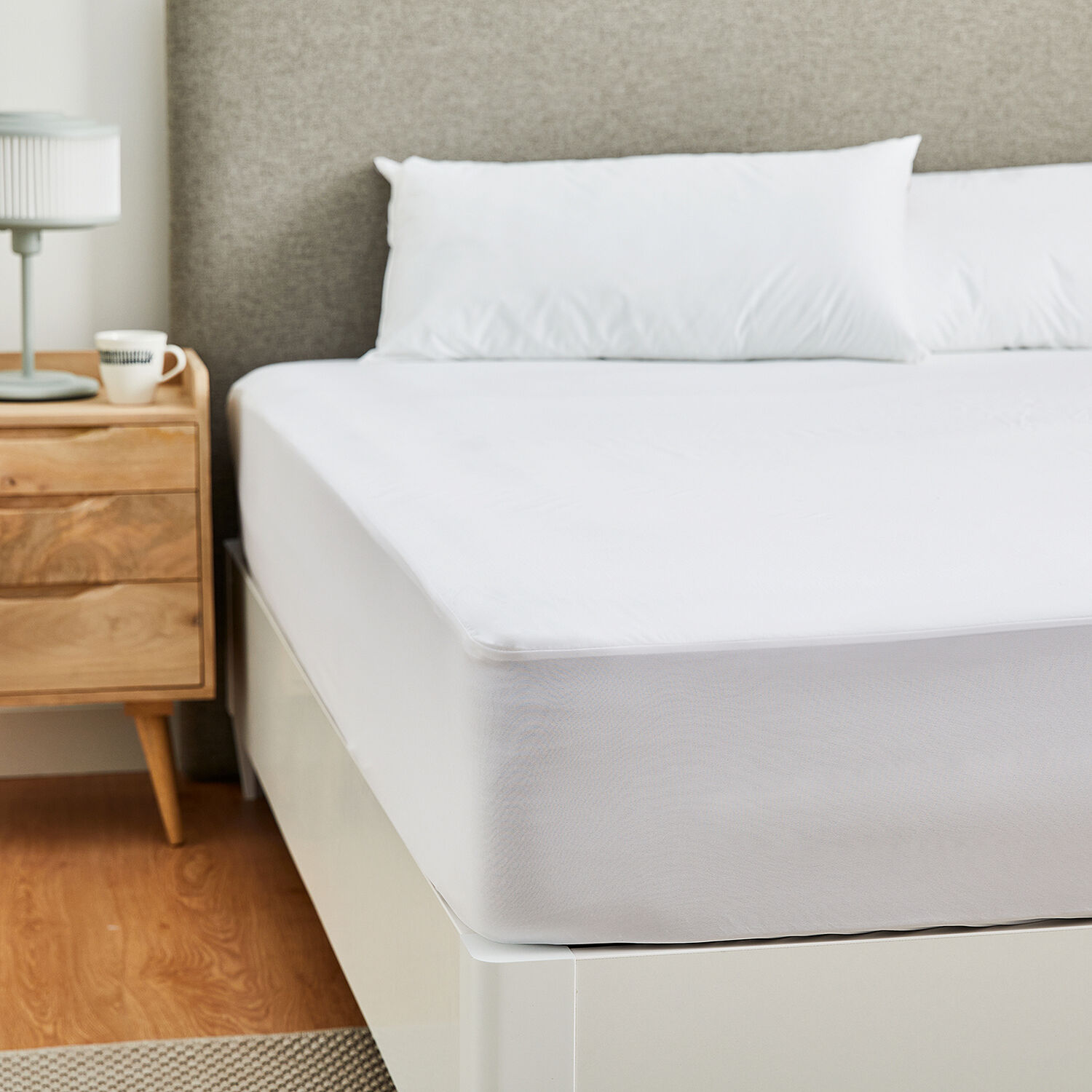 Una cama bien hecha con sábanas y almohadas blancas, una mesita de noche con lámpara y taza.