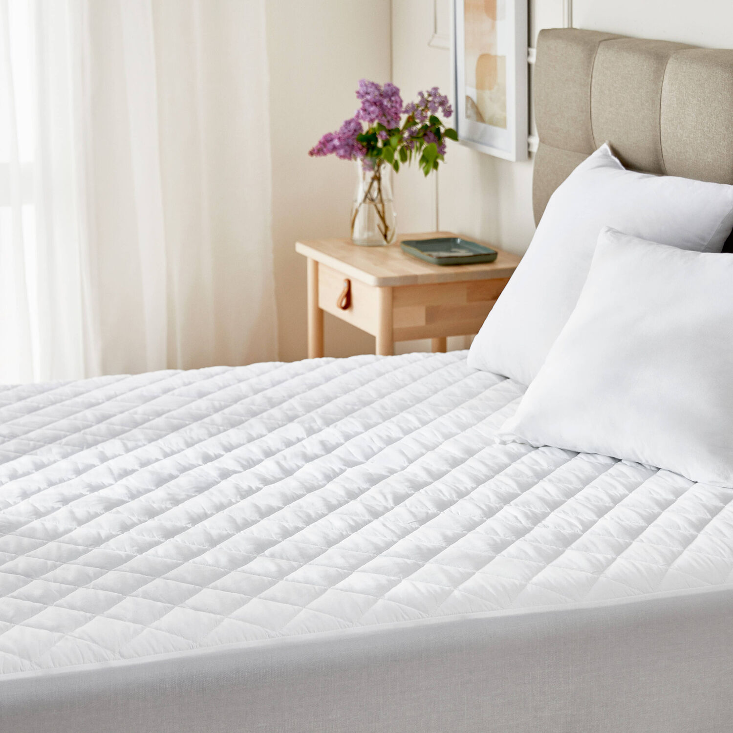 Un diván blanco acolchado con almohadas blancas y flores moradas en una mesita de noche.