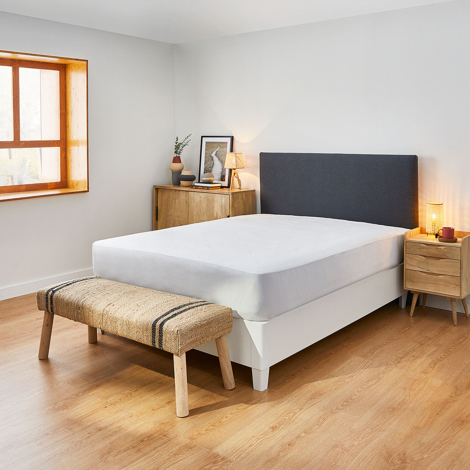 Una escena de dormitorio con cama blanca, cabecero gris, mesita de noche de madera y banco tejido.