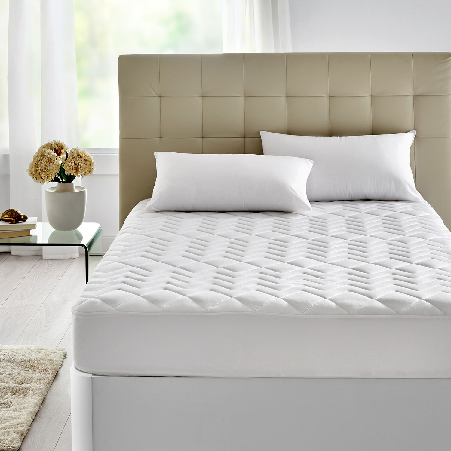Un sobrecolchón blanco acolchado sobre una cama con cabecero beige y almohadas blancas.