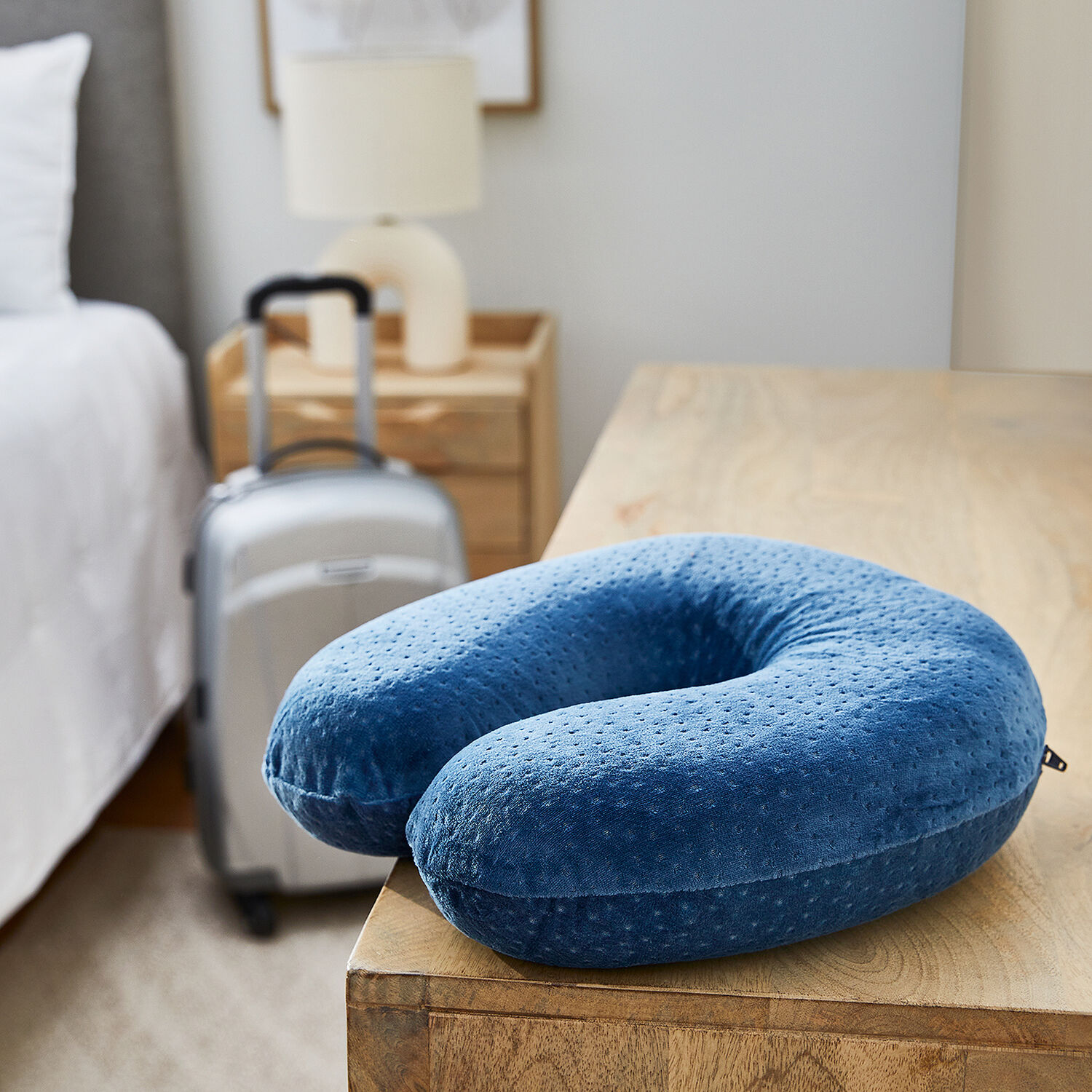 Una almohada de viaje azul descansa sobre madera, con maleta y cama al fondo.