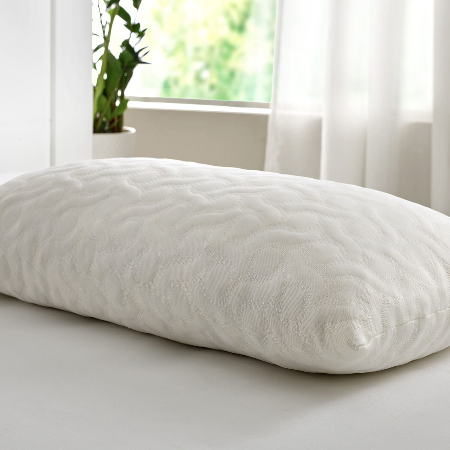Una almohada blanca estampada descansa sobre una cama blanca, con ventana y plantas al fondo.