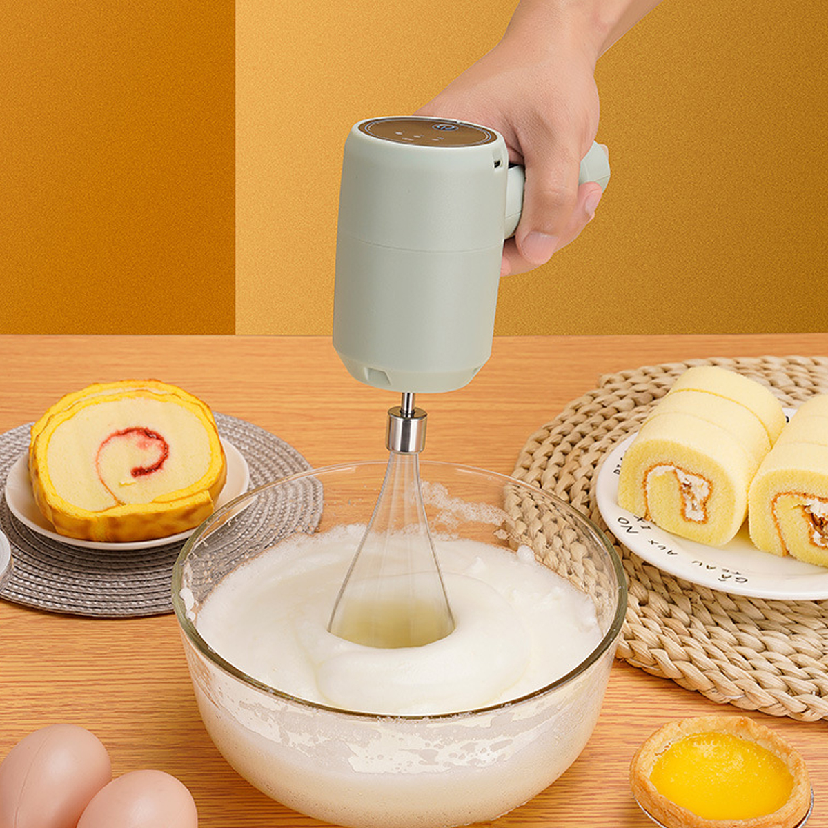 Handmixer schlägt Eiweiß in Schüssel; Kuchenrollen und Eier liegen daneben.