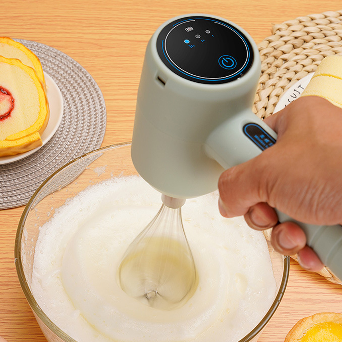 Eine Hand hält einen hellgrünen elektrischen Mixer und schlägt Eischnee in einer Glasschüssel. Kuchenstücke sind in der Nähe.