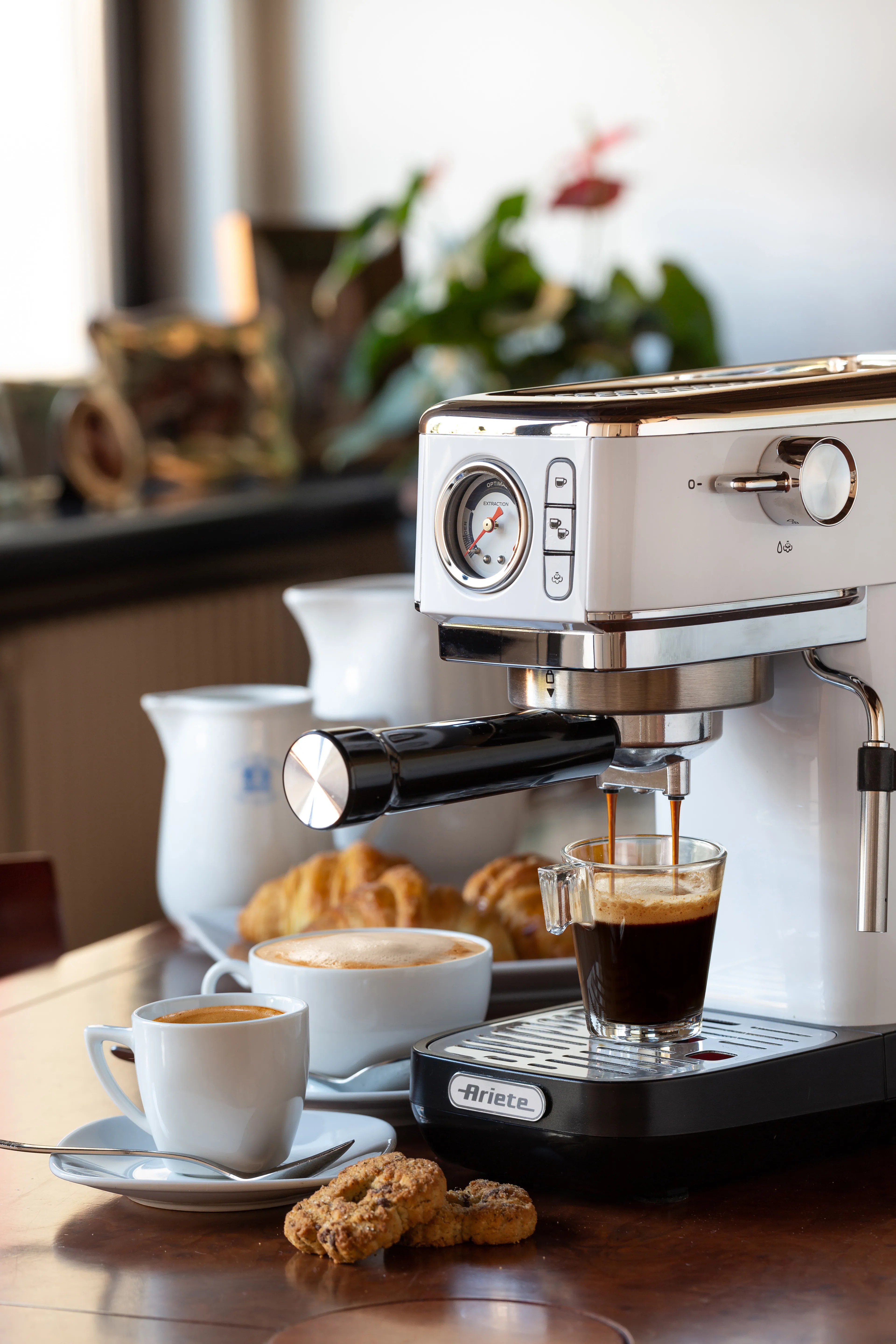 Eine weiße Espressomaschine brüht Kaffee in ein Glas, daneben Gebäck und Tassen.