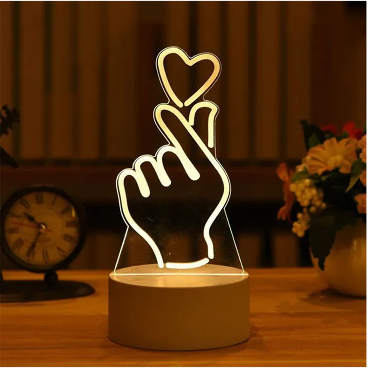 Eine Herz-Geste Lampe mit einem Herz oben, auf einem Holztisch mit Uhr und Blumen.